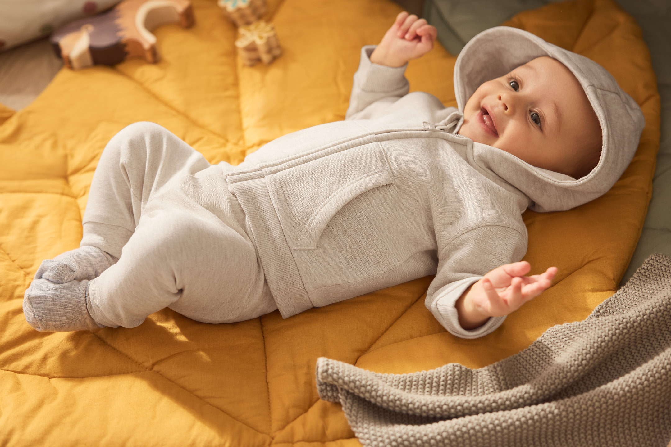 Happy baby in a grey hooded outfit lying on a yellow quilted blanket with wooden toys.