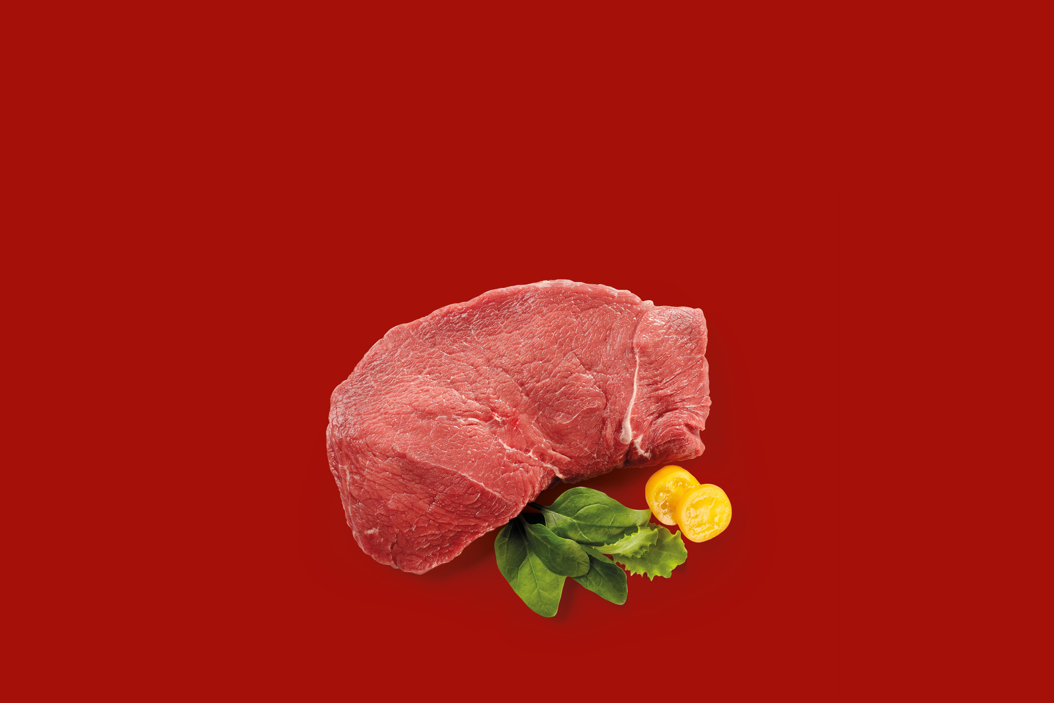 Raw beef steak with fresh spinach leaves and yellow cherry tomatoes on a red background.