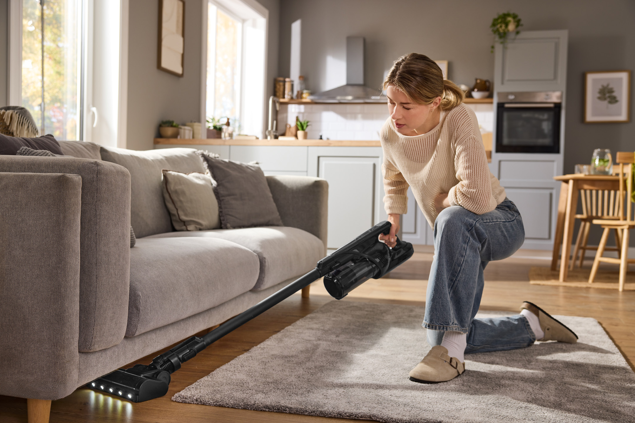 Woman vacuuming under a sofa with a cordless stick vacuum cleaner in a modern living room.