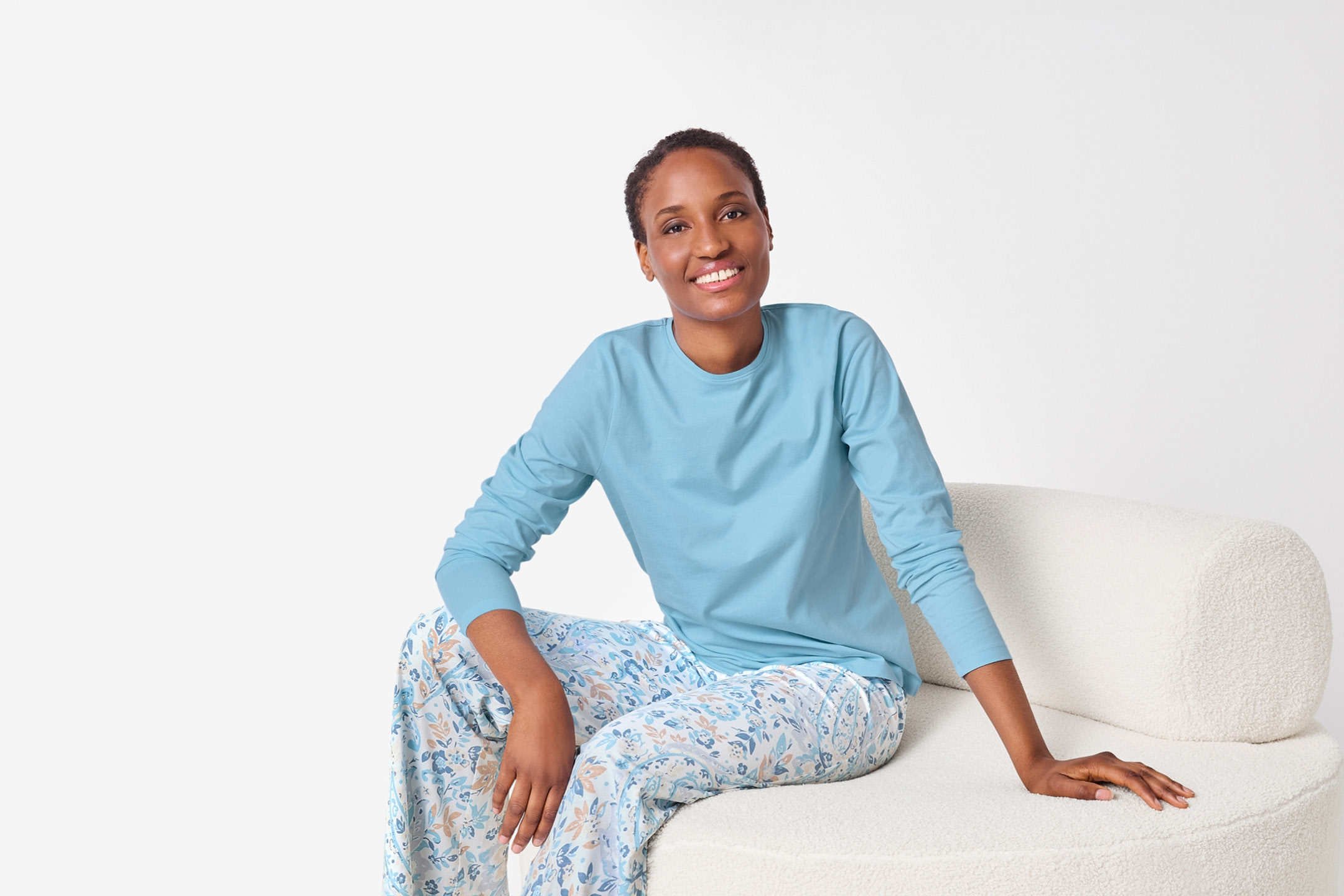 Woman in blue long-sleeve top and floral pyjama bottoms, sitting on a white sofa.