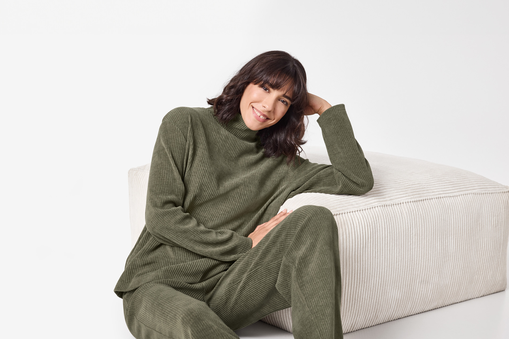Woman in a green ribbed loungewear set, sitting on a white pouffe, smiling at the camera.