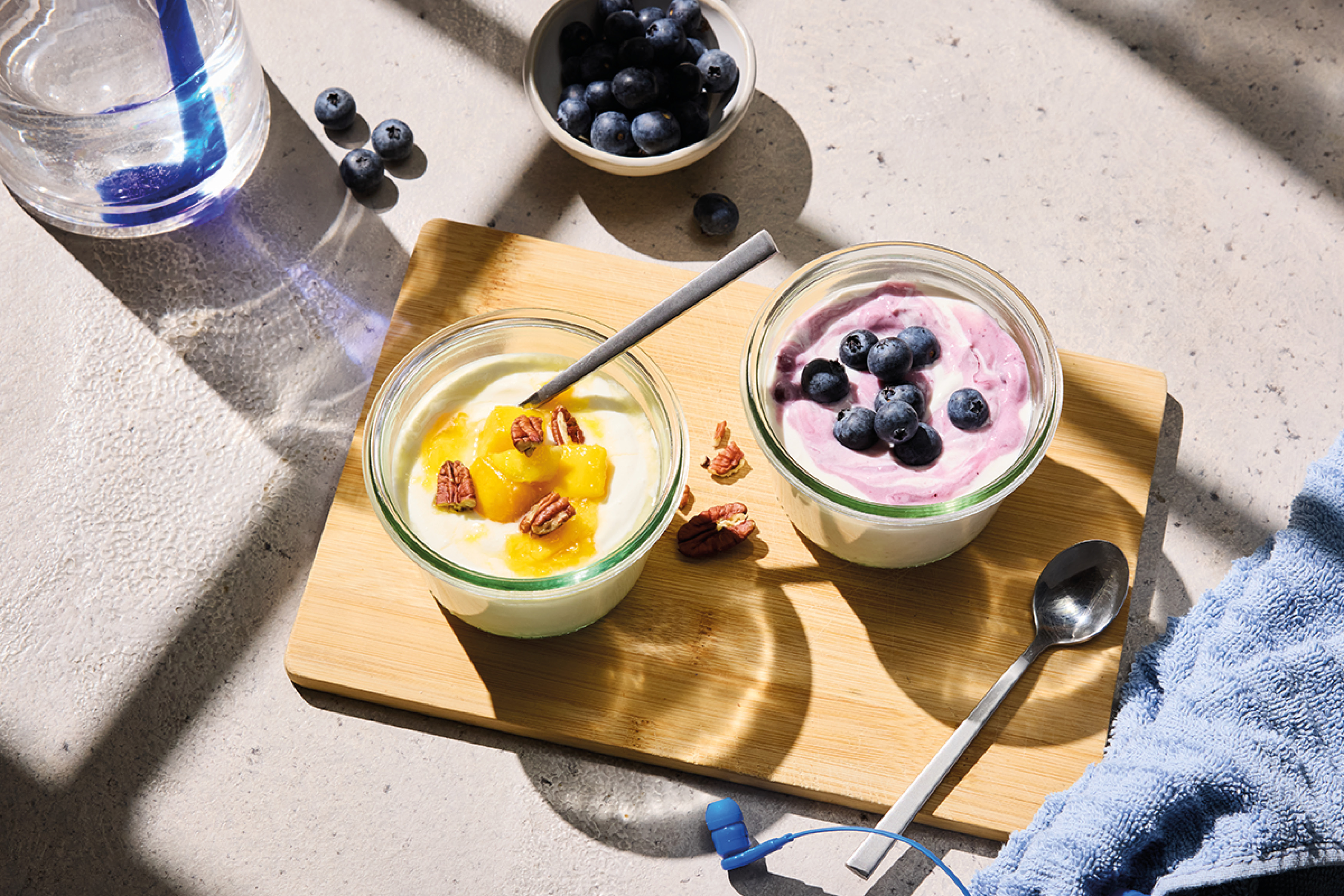 Two yogurt bowls with fruit and nuts on a wooden board, with blueberries and water.