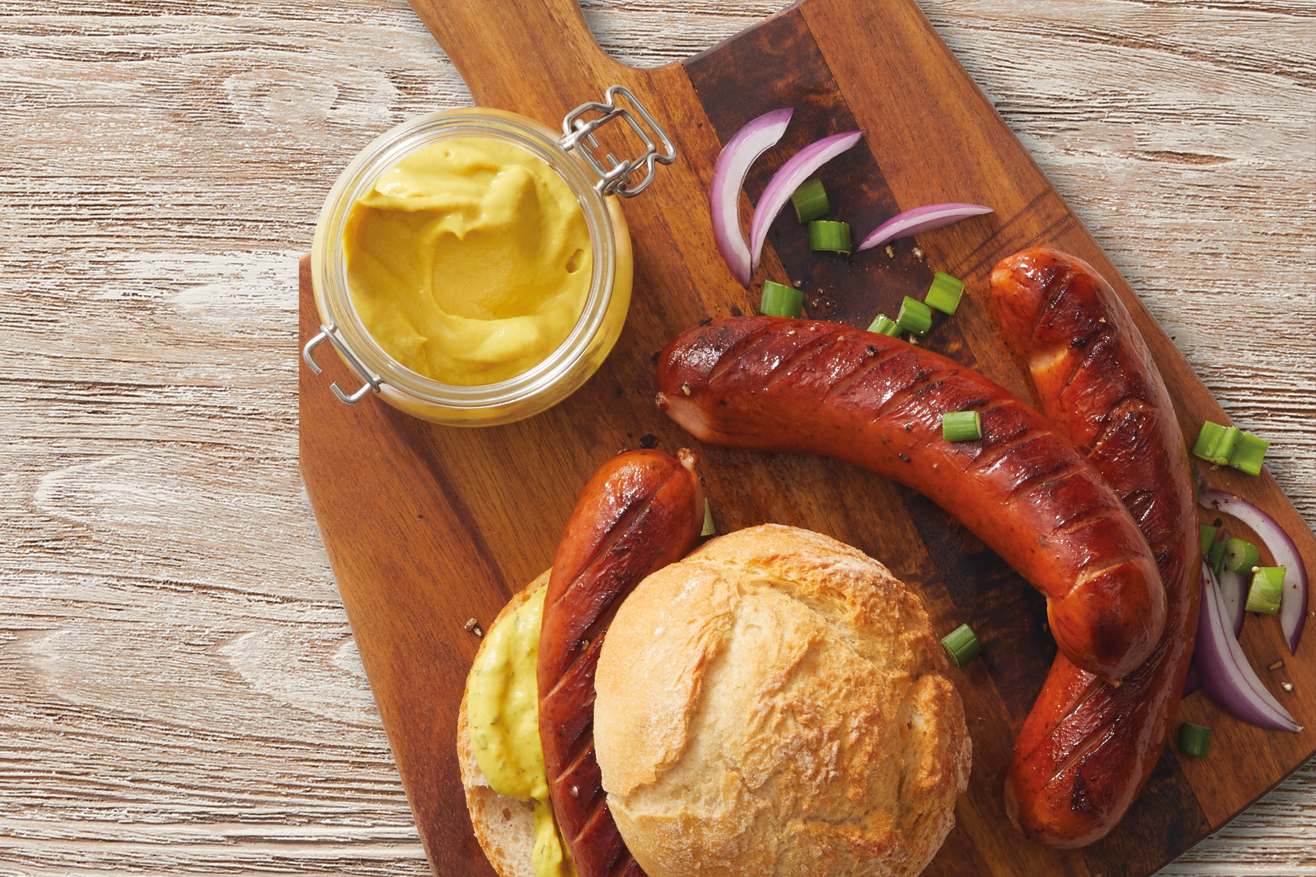 Grilled sausages with mustard, red onion, and spring onion on a wooden board.
