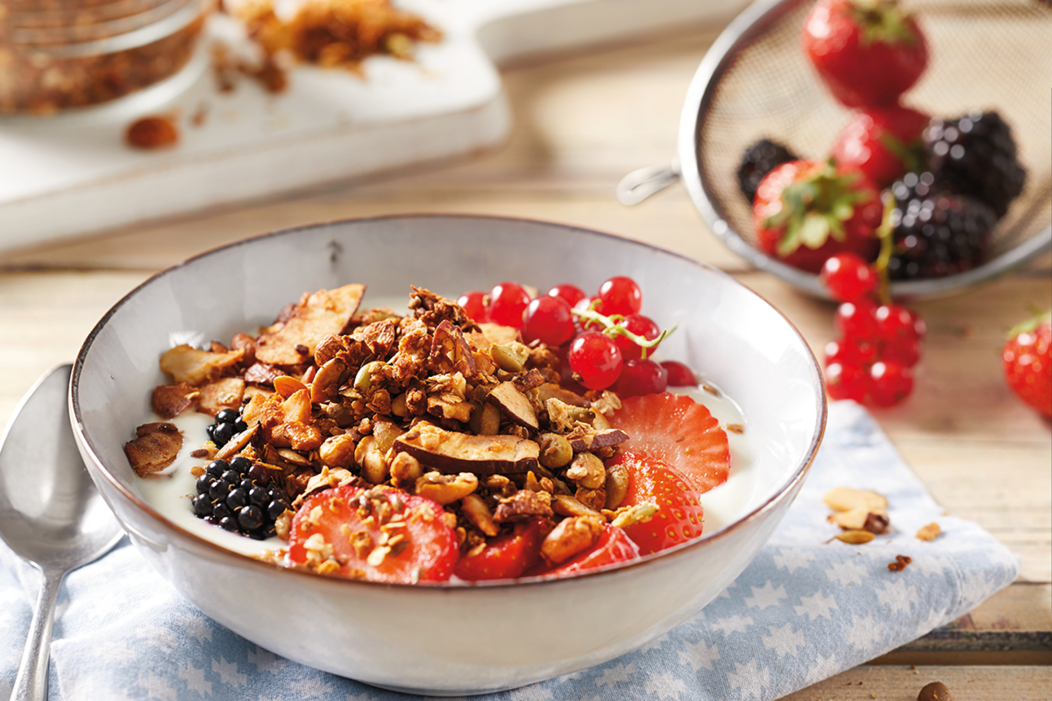 Granola with yogurt and fresh berries in a bowl, with a sieve of berries in the background.