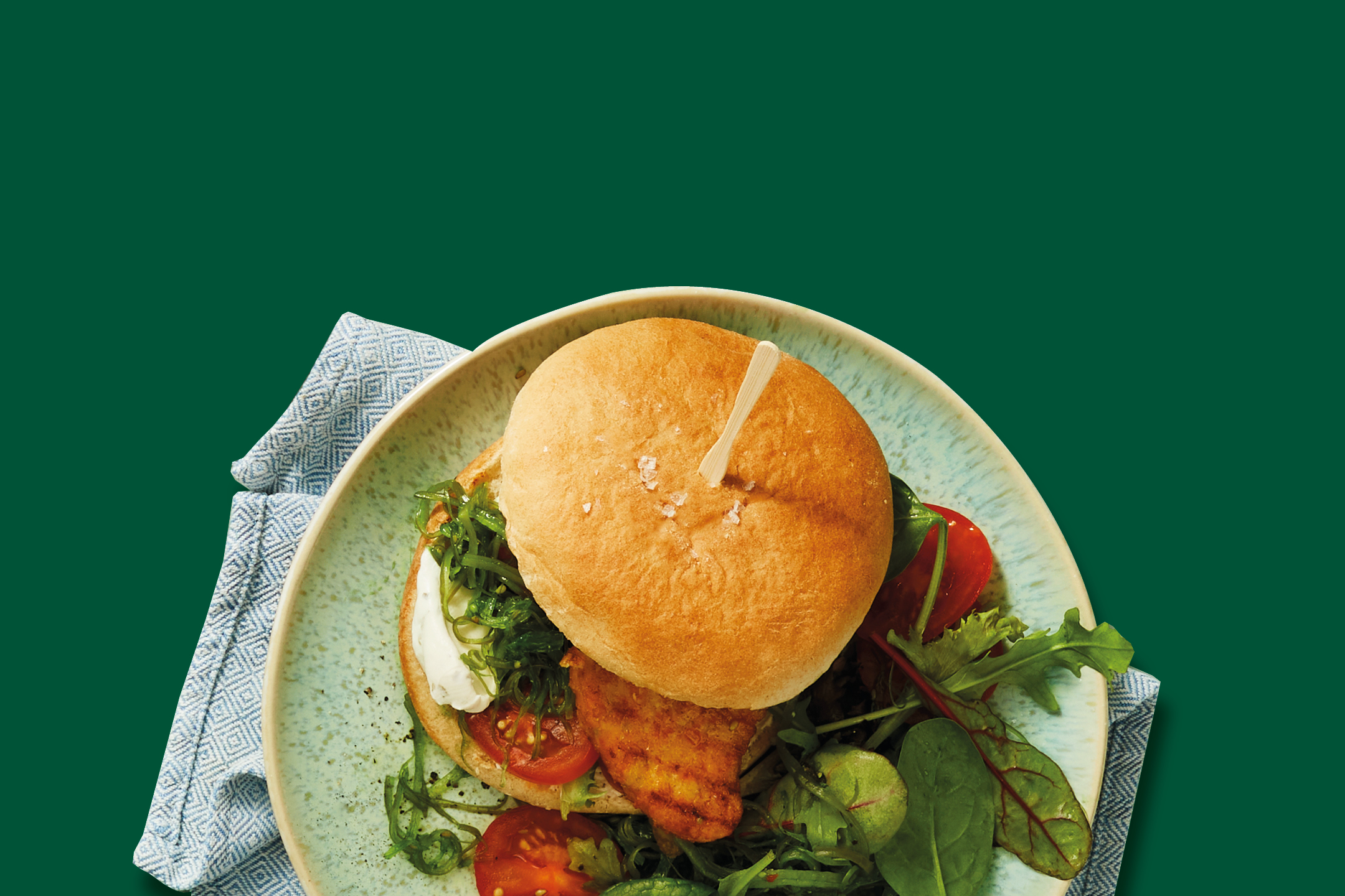 Top-down view of a burger with chicken, tomatoes, and greens on a plate with a napkin.