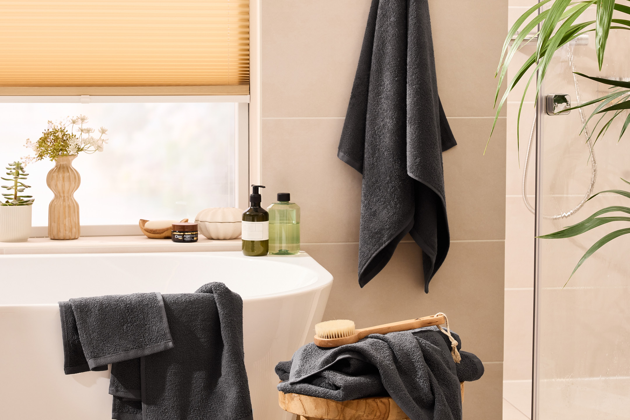 Bathroom with dark grey towels, bath, and shower, featuring plants and bath products.