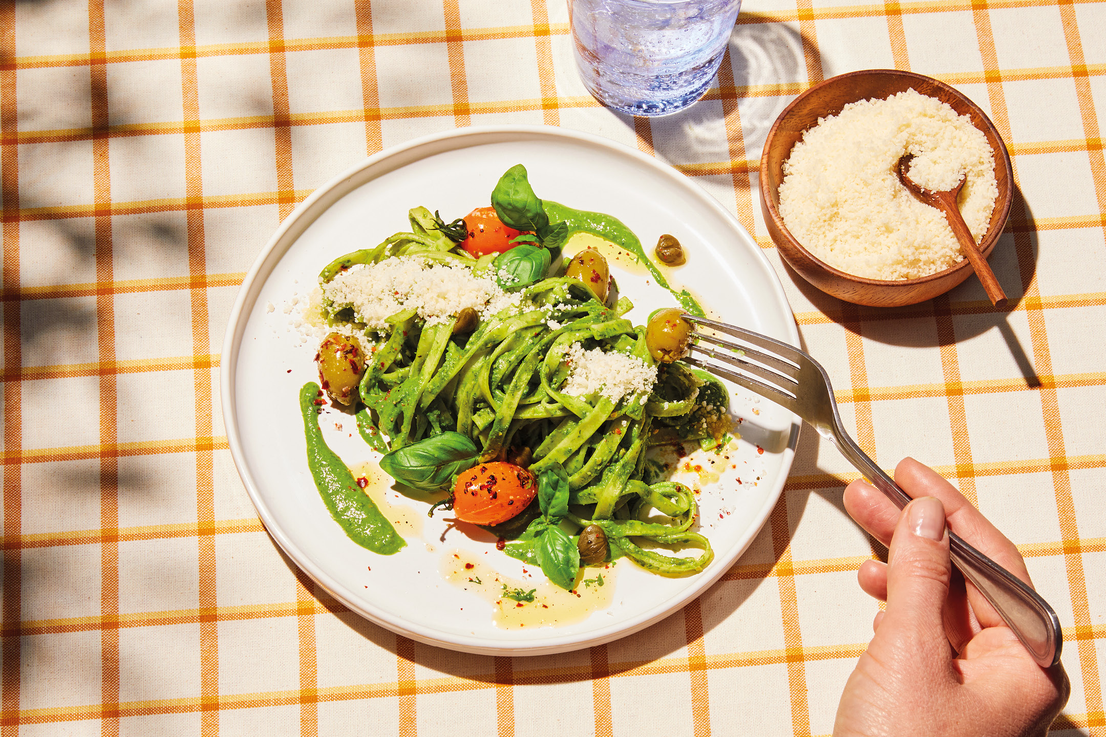 Green pasta with tomatoes, olives, basil, and grated cheese on a checkered tablecloth.