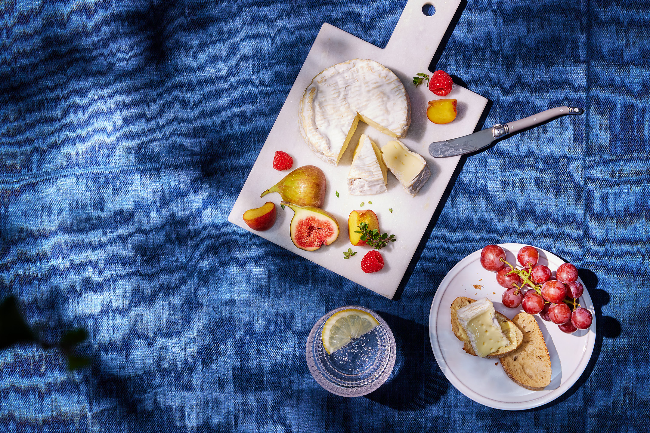 Cheese board with brie, figs, raspberries, and peaches on a blue tablecloth.