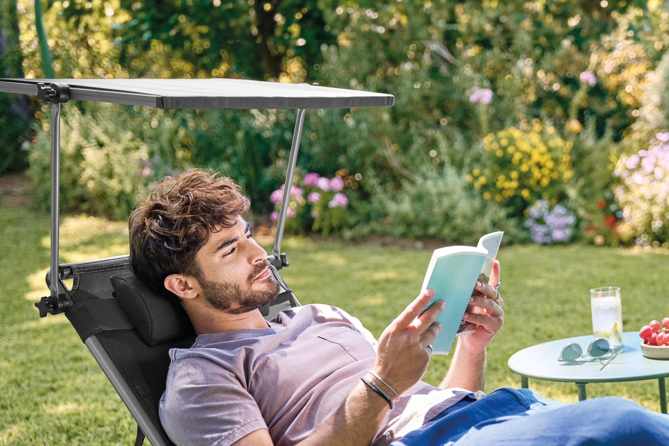 Man relaxing in a sun lounger with a sunshade, reading a book in a garden.