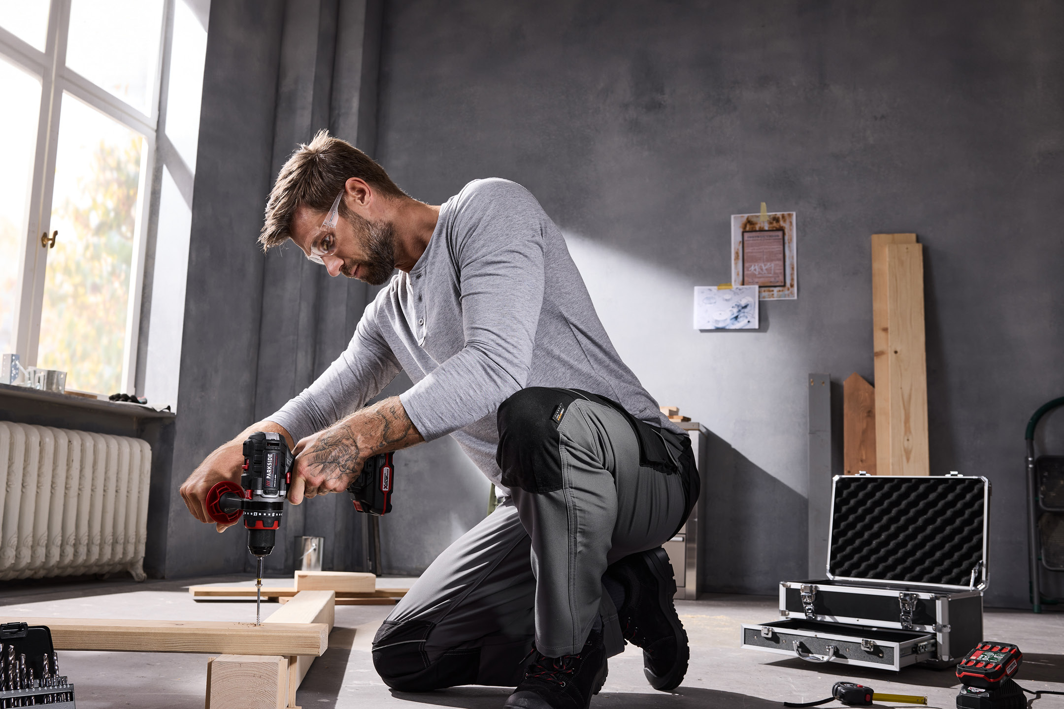 Man in workwear using a cordless drill to screw wood, with a tool case nearby.
