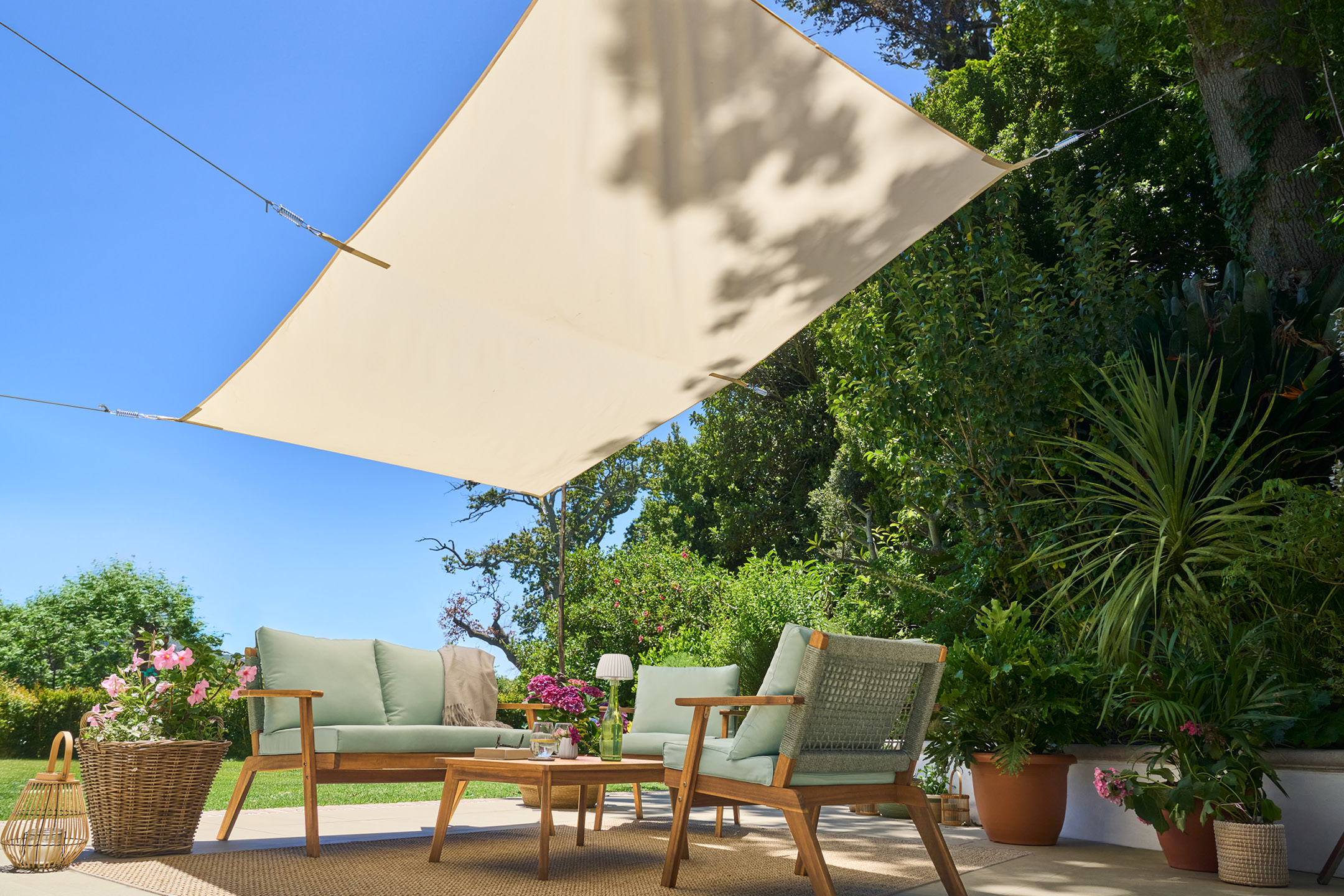 Outdoor patio with wooden sofa and armchairs with light green cushions under a sun shade.