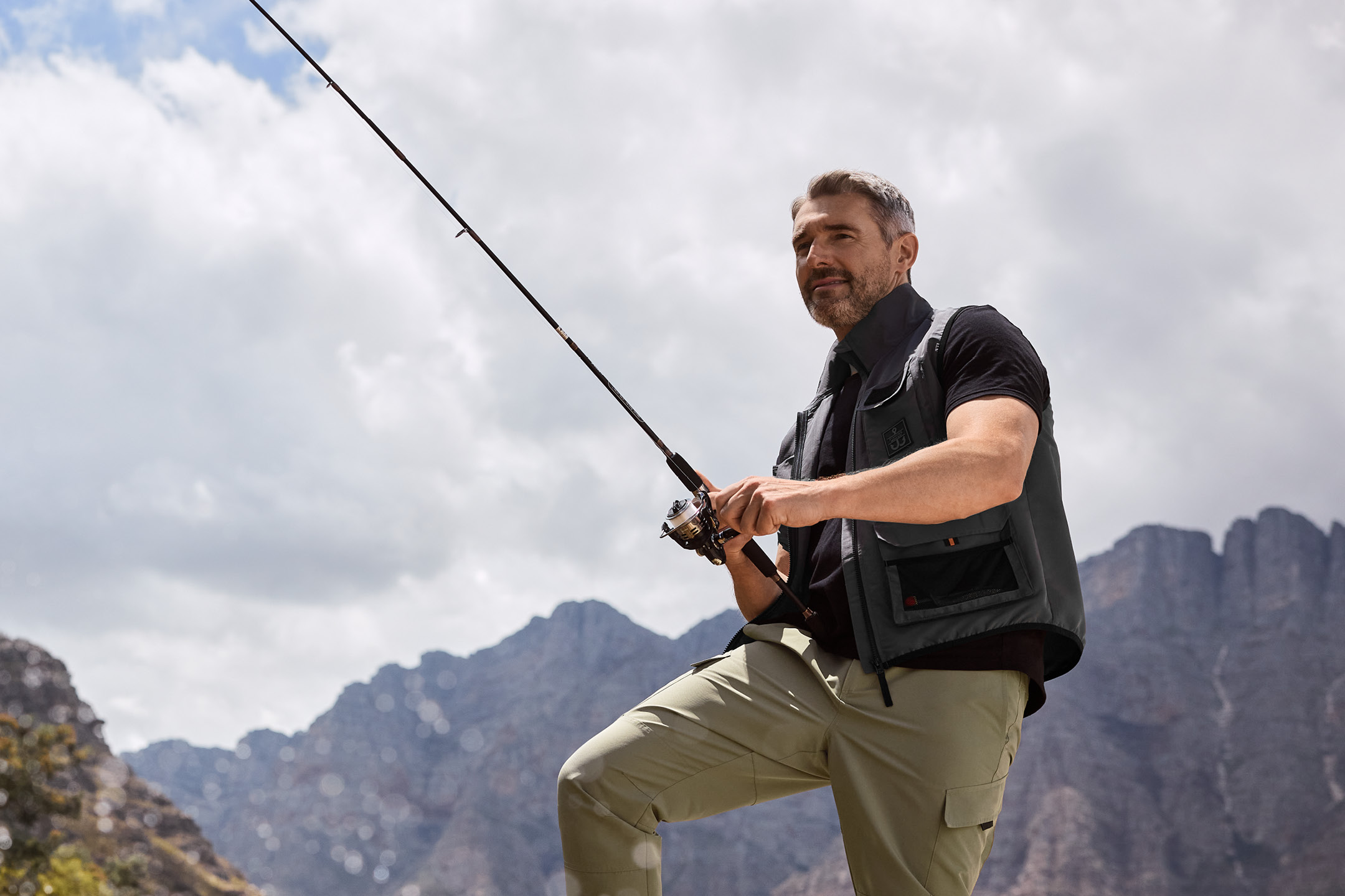 Man in a fishing vest and cargo pants holding a fishing rod, with mountains in the background.