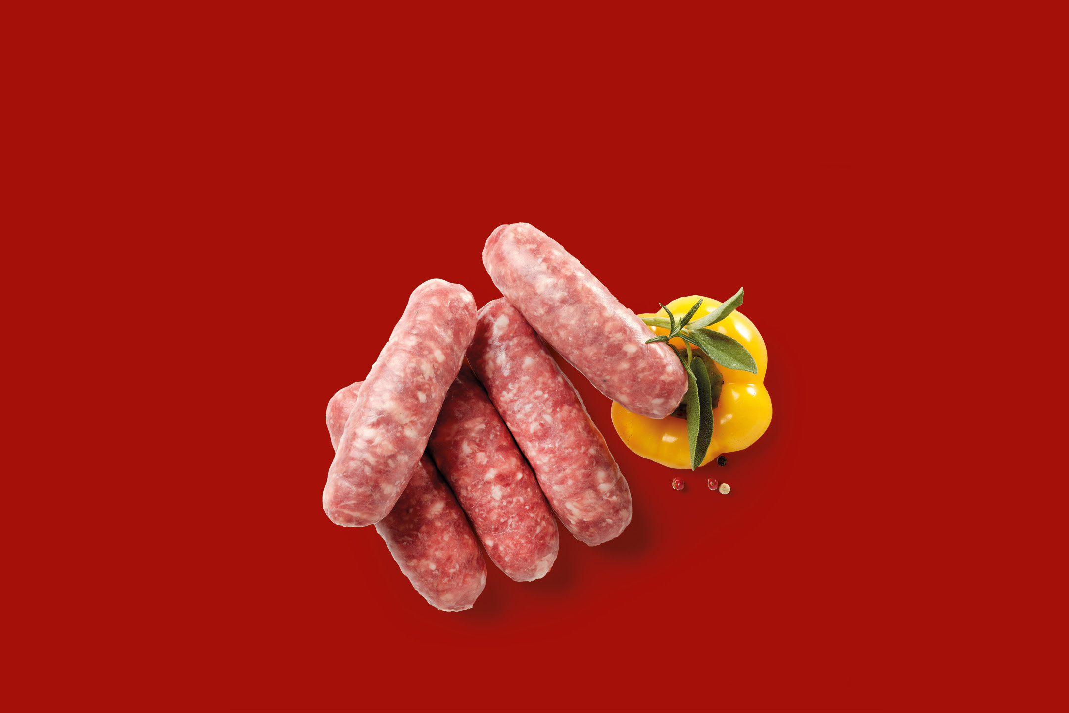 Raw sausages with a yellow bell pepper and herbs on a red background.