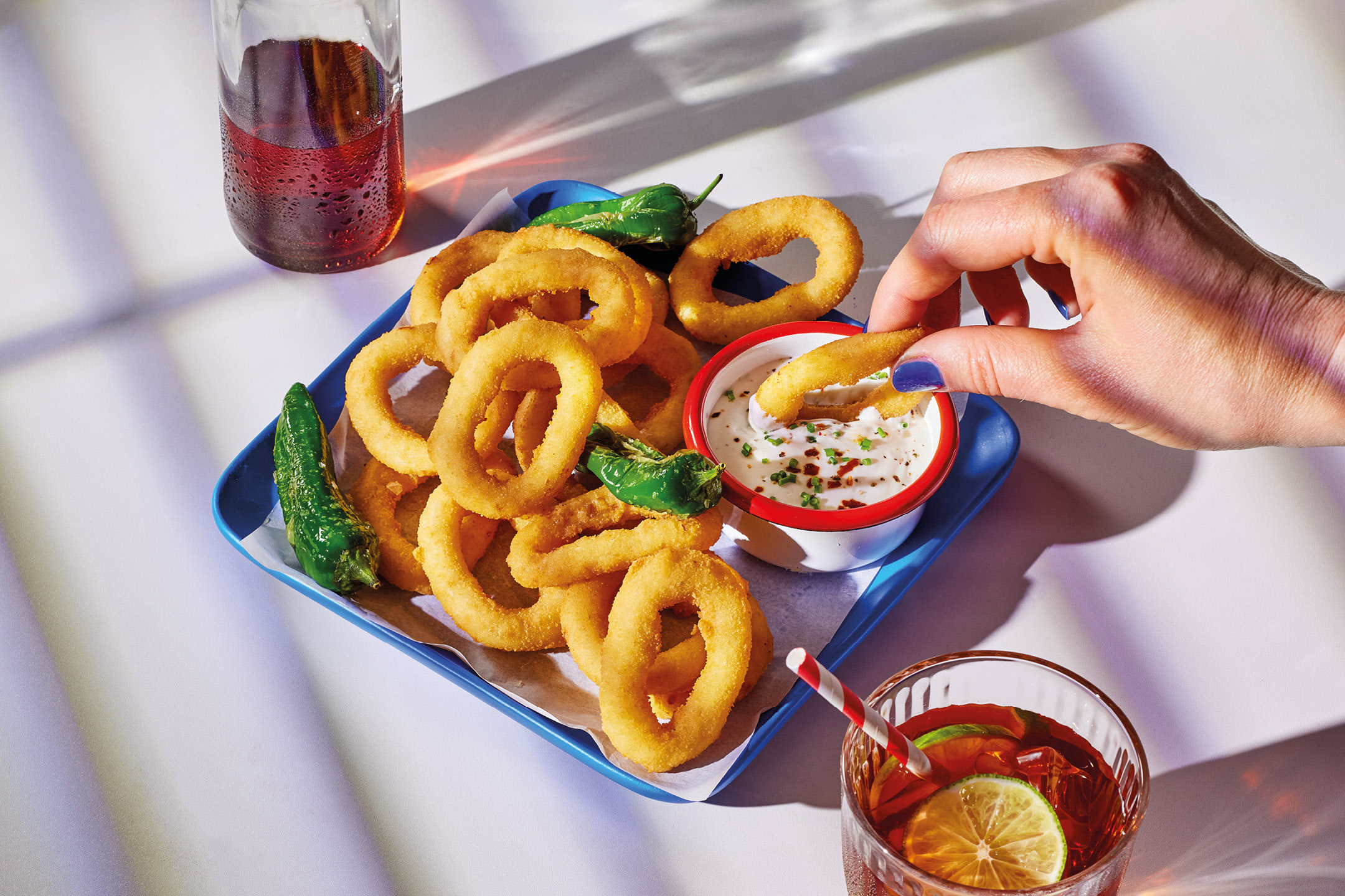 A hand dips a golden onion ring into a white sauce, with a drink nearby.