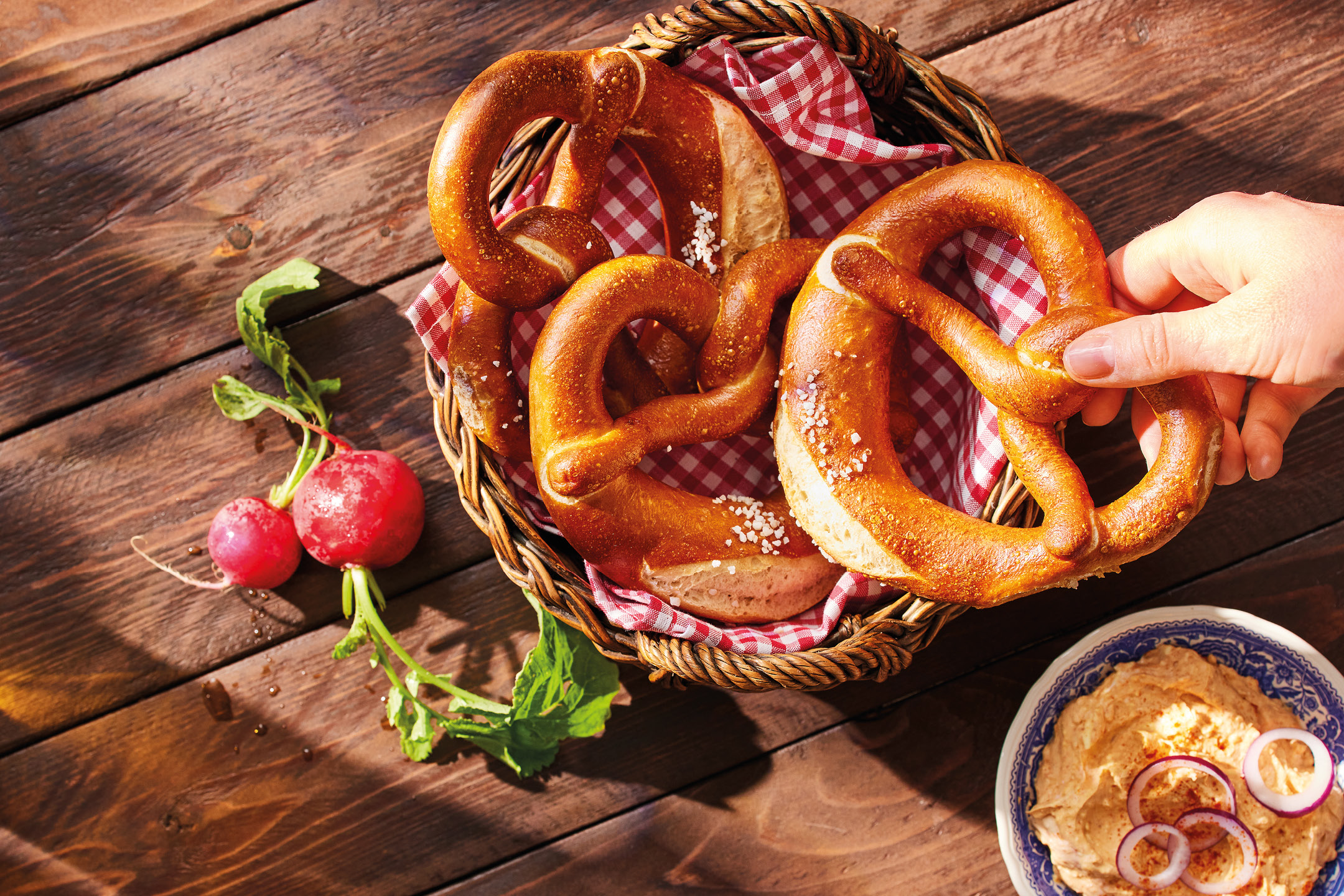 A hand reaching for a pretzel from a basket, with radishes and a dip on a wooden table.