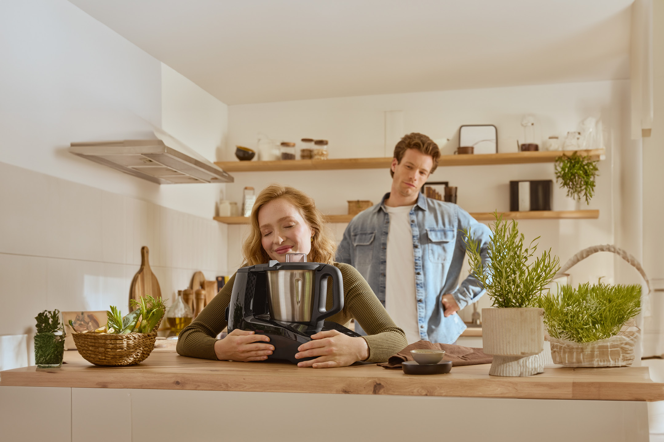 A woman happily embracing a kitchen machine on a counter, with a man looking on in the background.