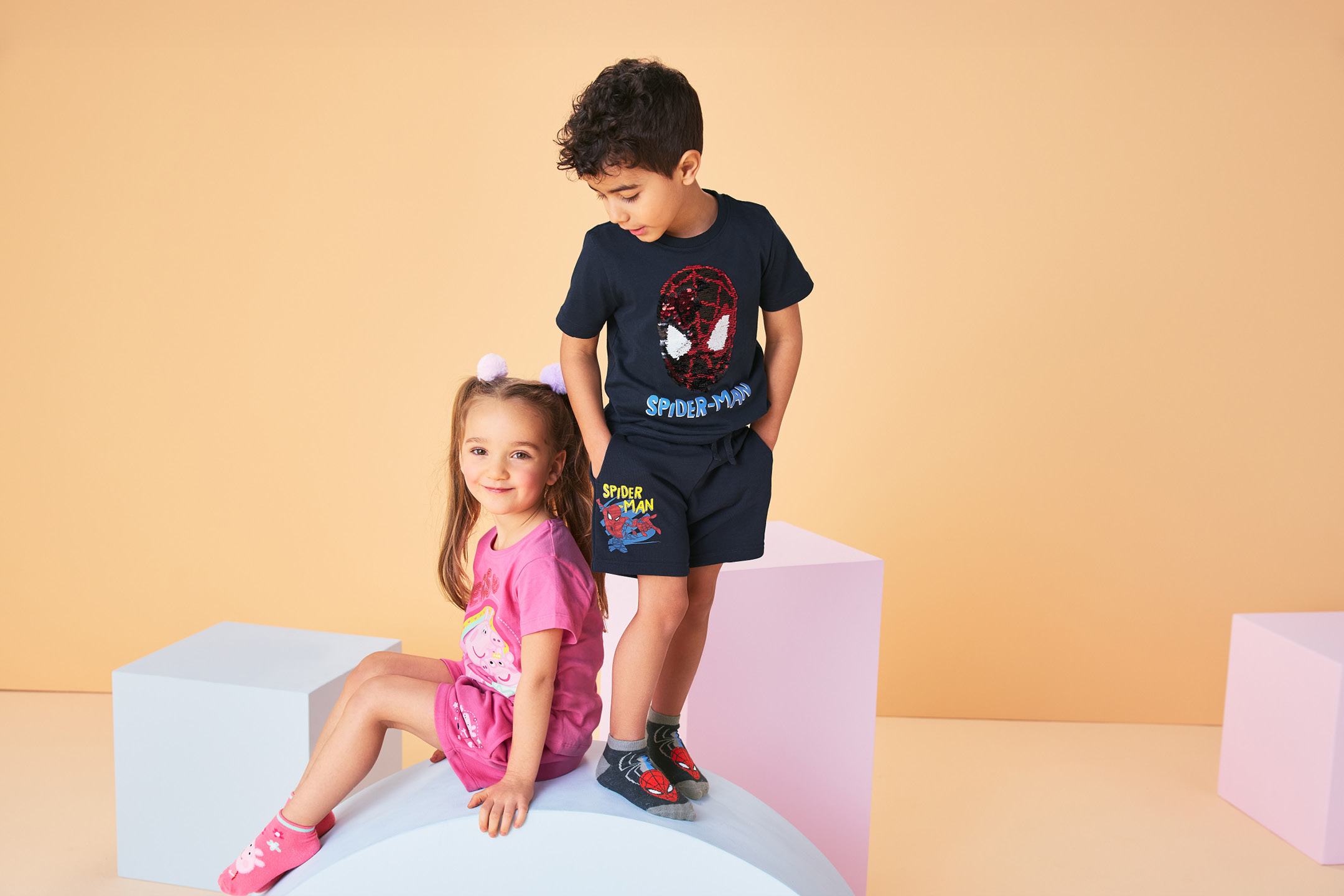 Two children wearing character-themed t-shirts and shorts, standing and sitting on blocks.