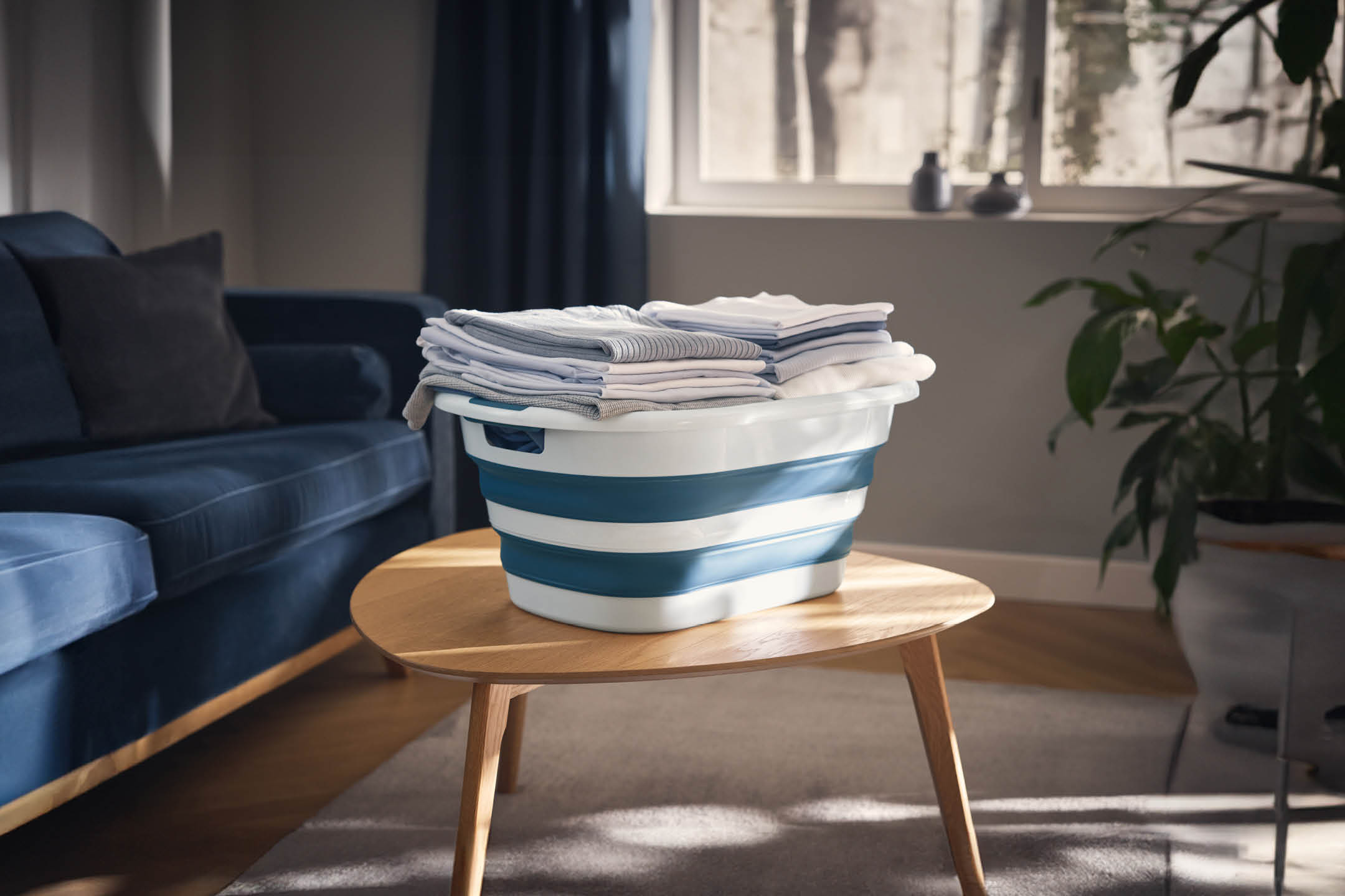 Foldable laundry basket with neatly folded clothes on a wooden coffee table in a living room.