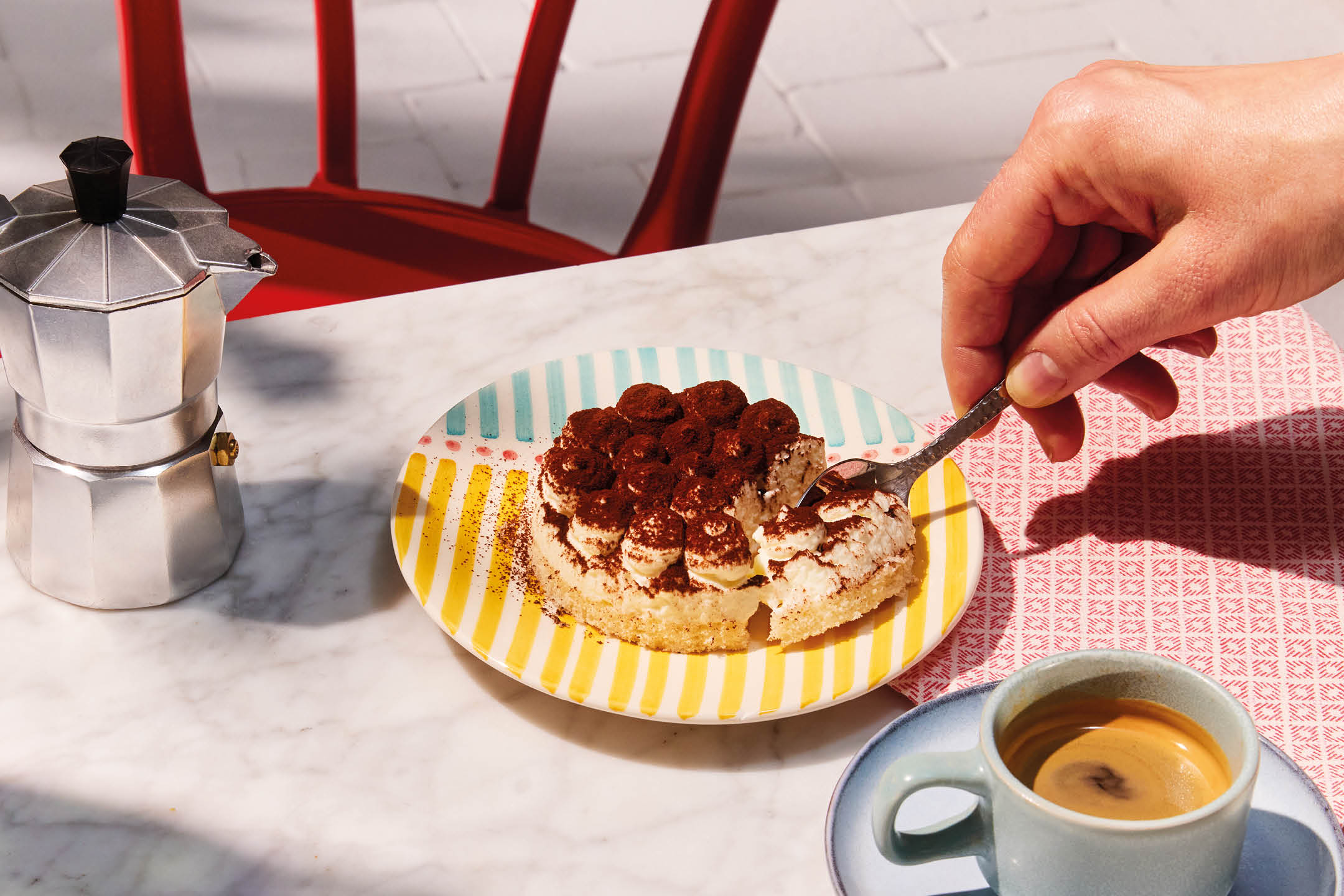A hand spoons tiramisu from a colorful plate next to a moka pot and a cup of espresso.