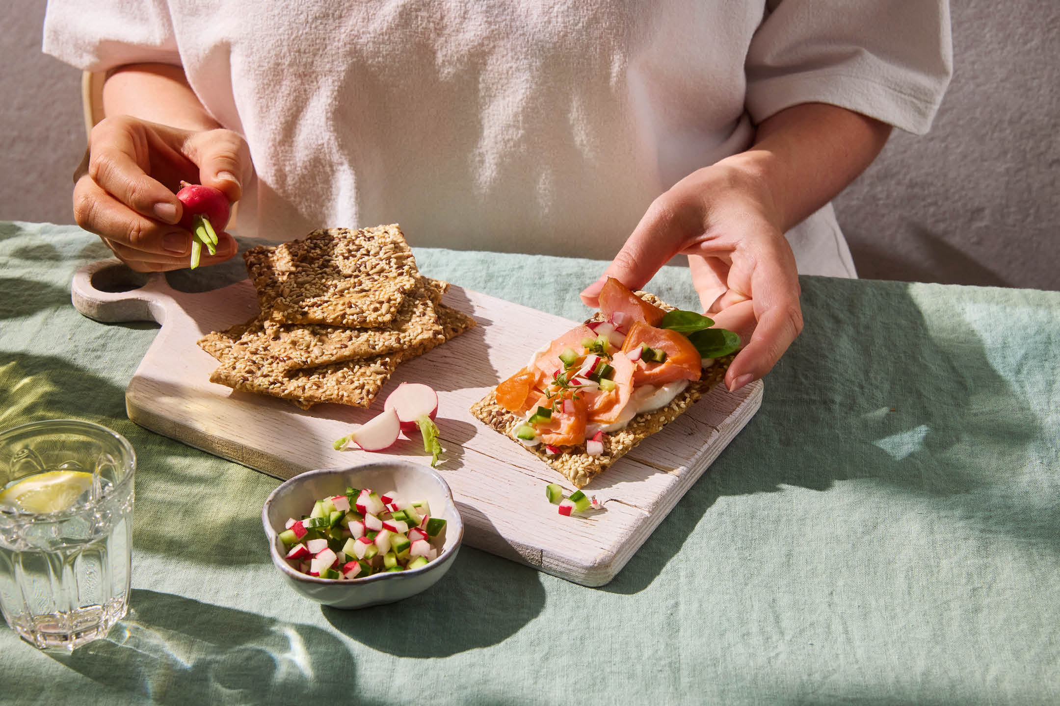 Hands preparing a crispbread snack with smoked salmon, cream cheese, and diced radishes.