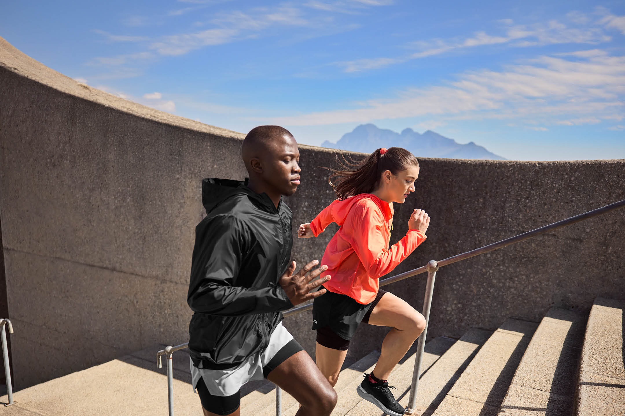 Two athletes in activewear running up outdoor stairs, with mountains in the background.