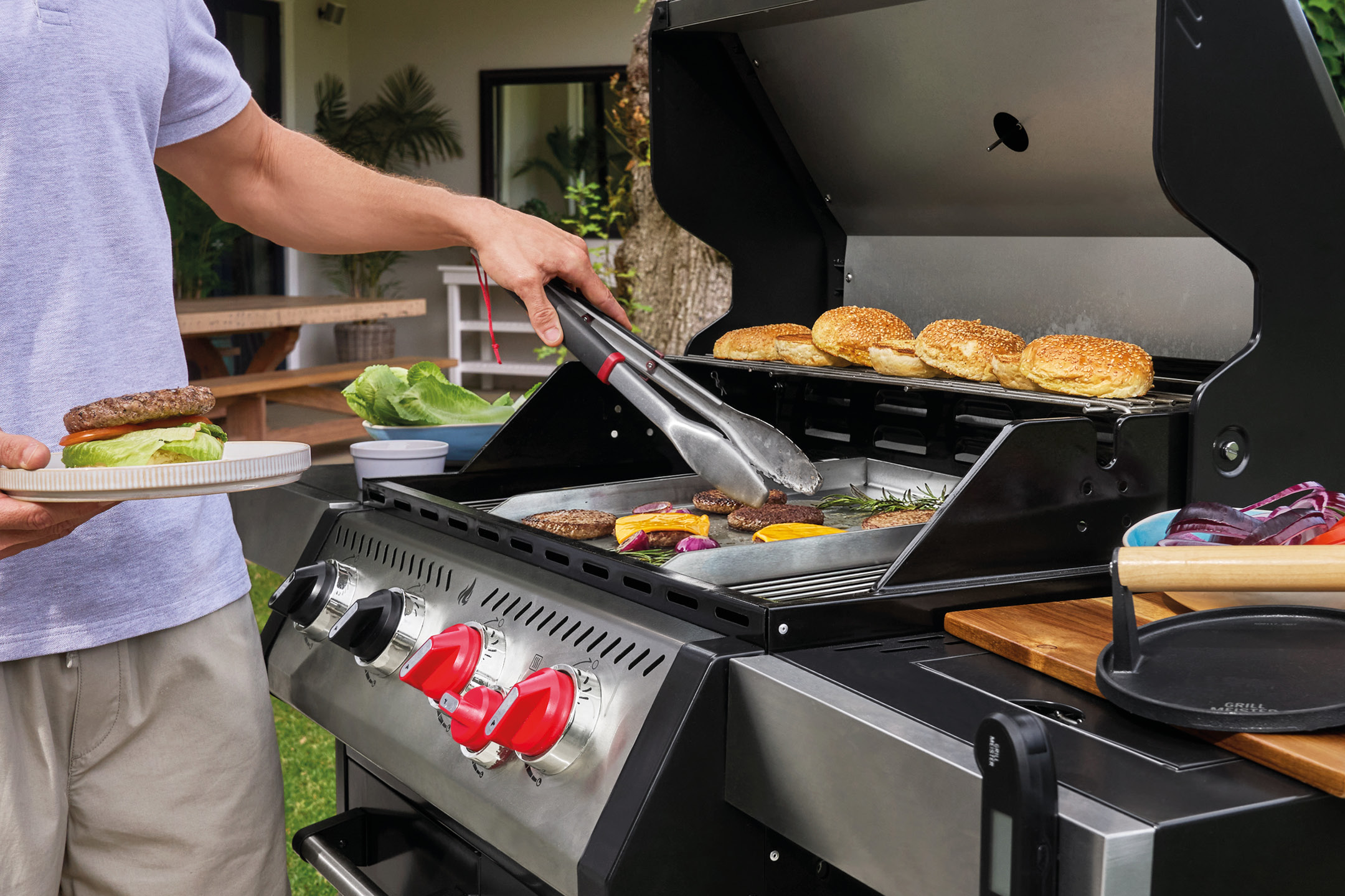 Man grilling burgers and buns on an outdoor gas grill, holding a finished burger.