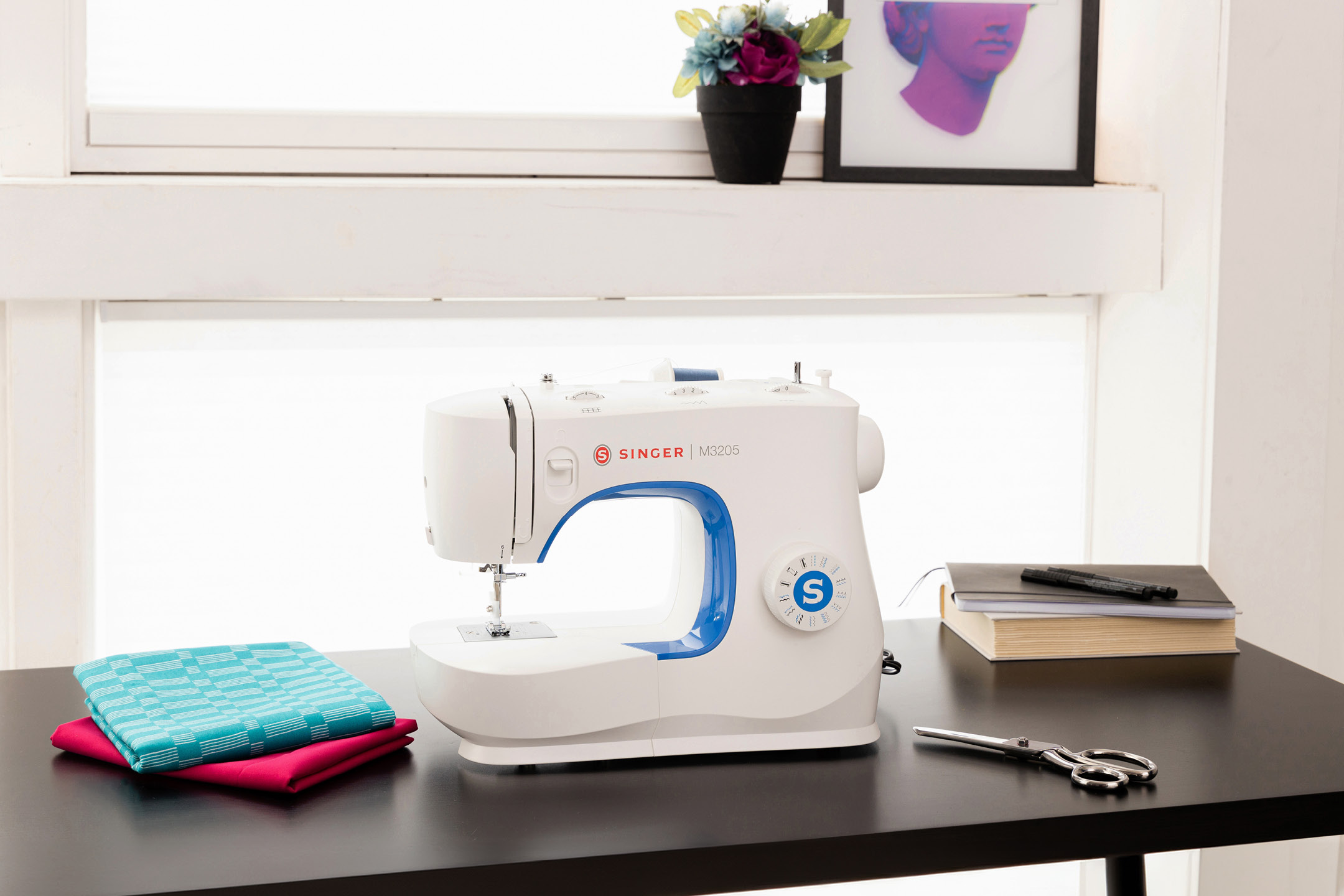 White sewing machine with blue accents on a dark table with folded fabrics, scissors, and notebooks.