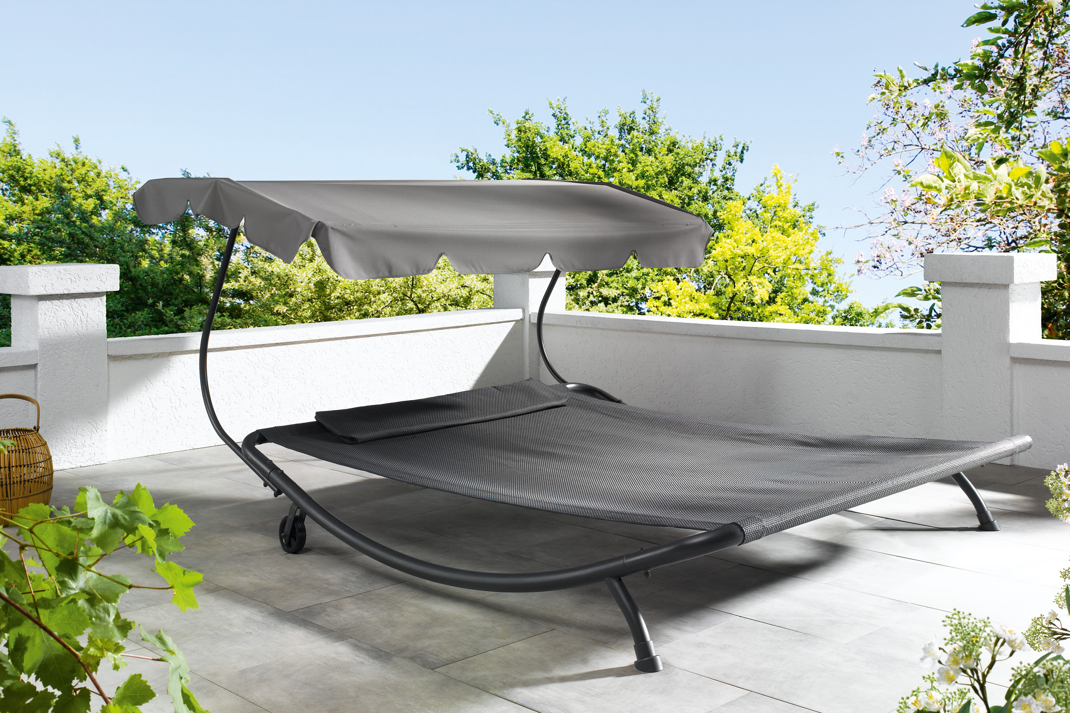 Grey sun lounger with canopy and pillow on a balcony with green trees in the background.