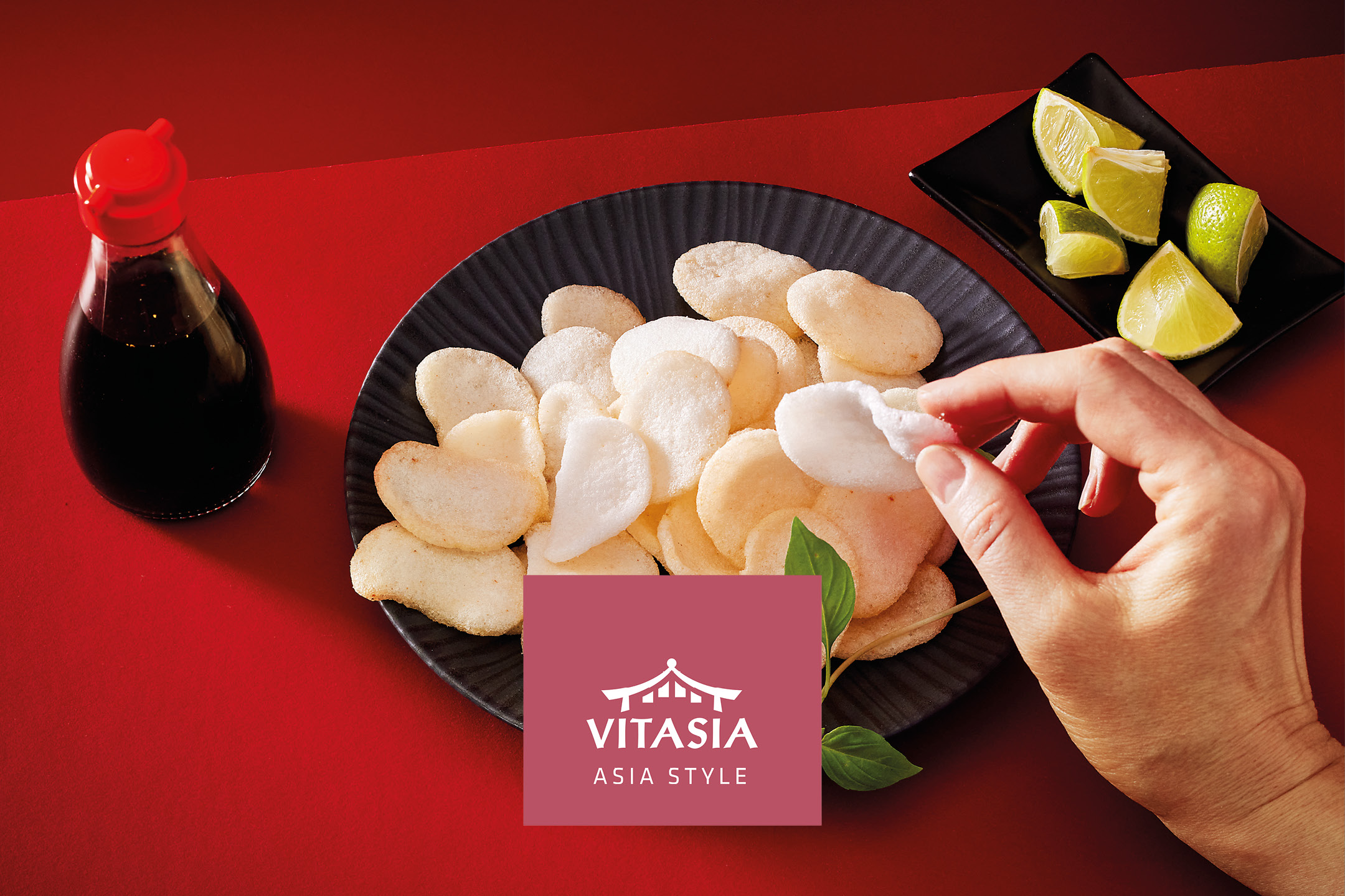 Prawn crackers on a black plate with soy sauce, lime wedges, and a hand picking one up.