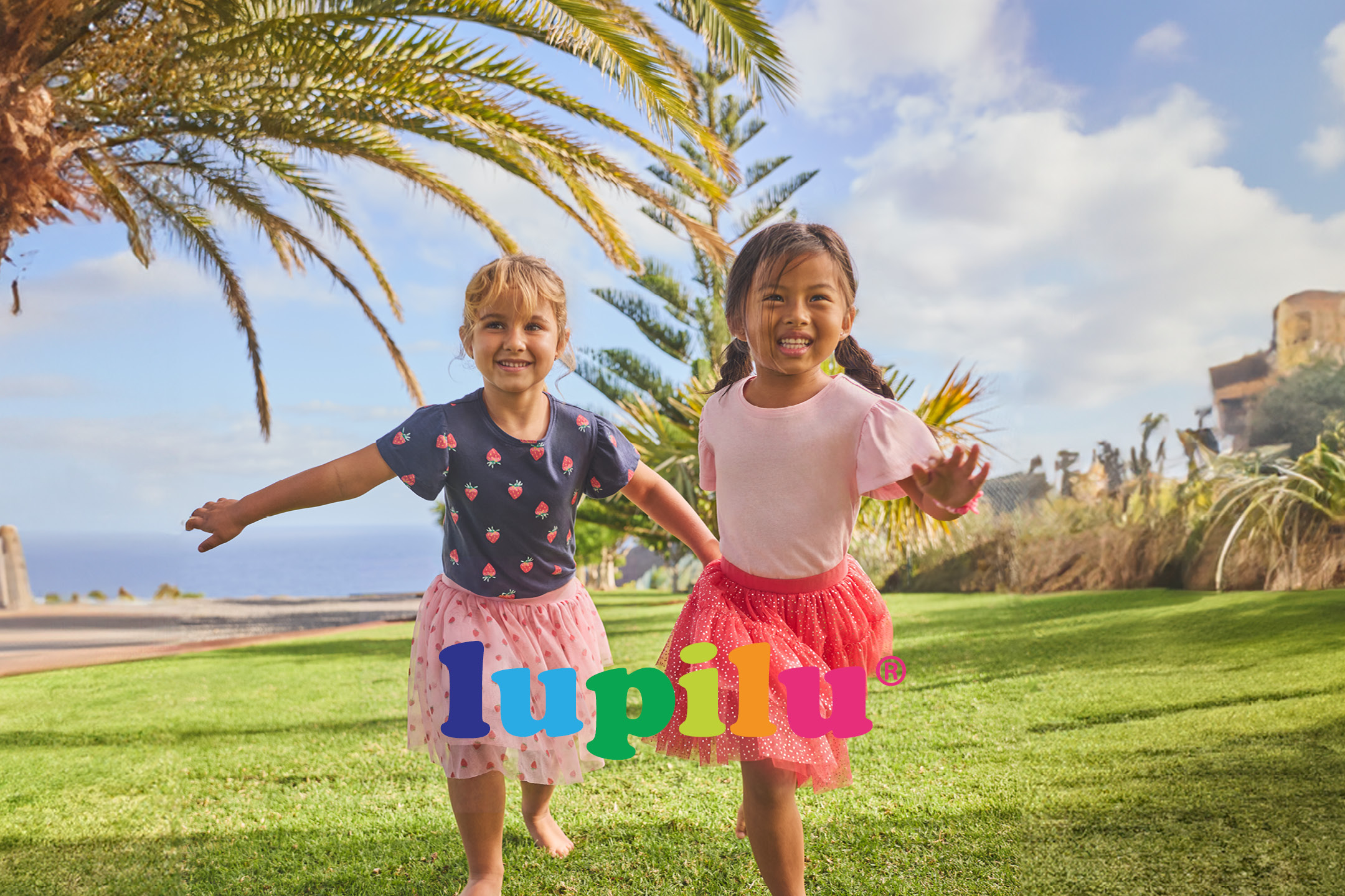 Two girls in summer clothes and skirts playing on grass with palm trees and ocean.