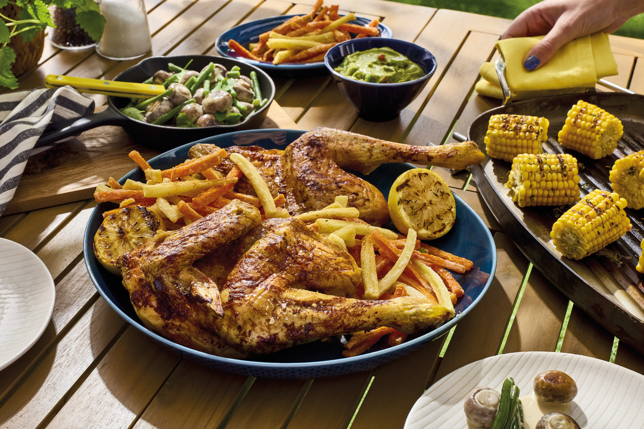 Grilled chicken with fries, corn on the cob, and vegetables on a wooden table.