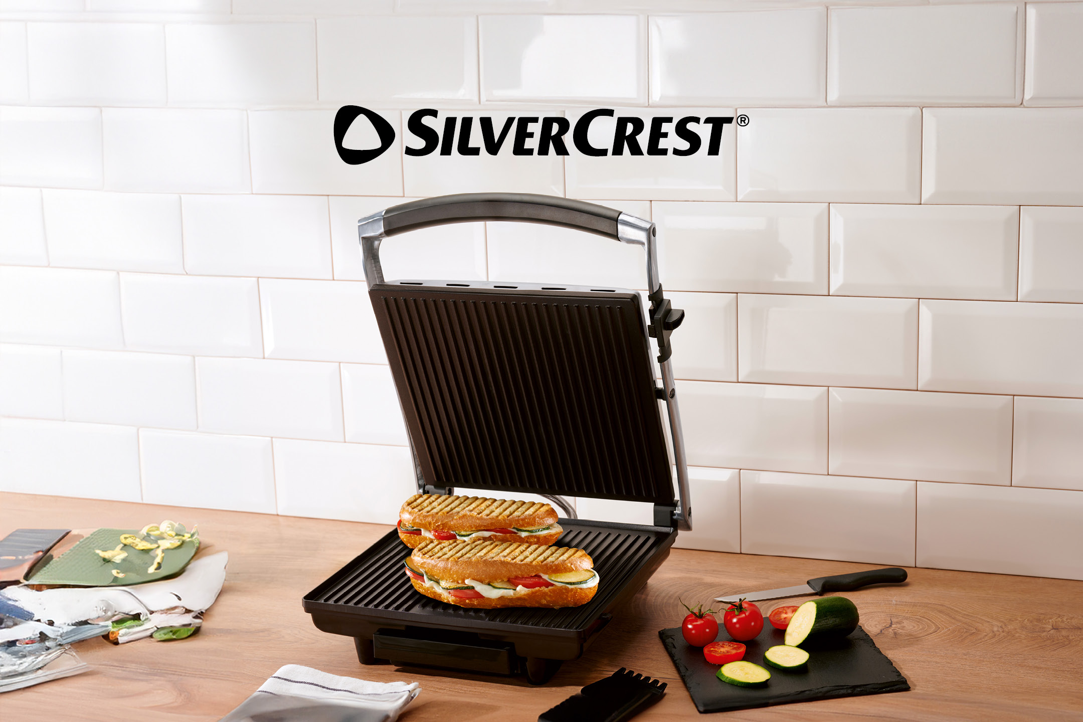 Open grill with two toasted sandwiches, vegetables, and kitchen utensils on a wooden counter.