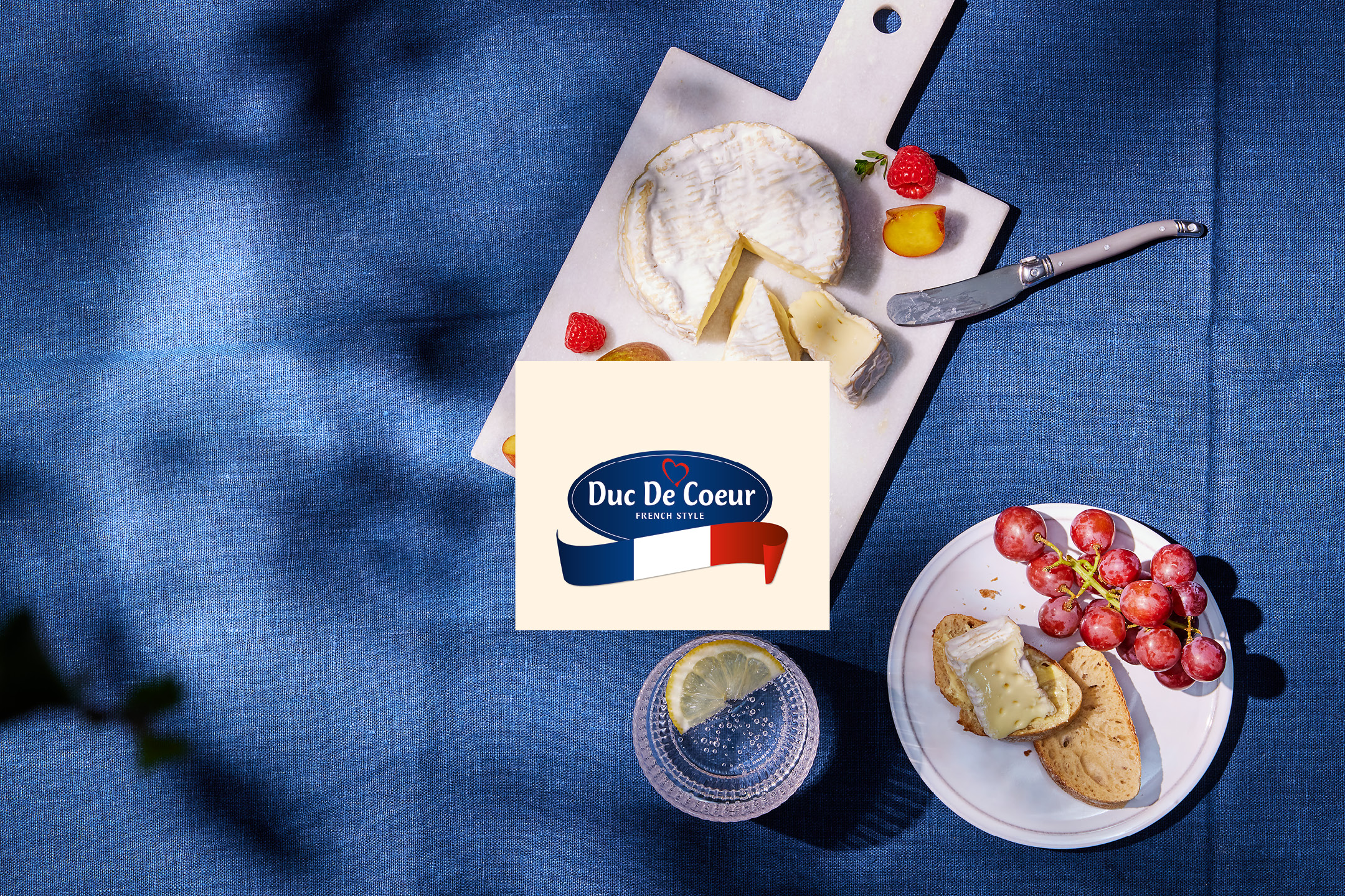 French cheese, bread, and grapes on a blue tablecloth, with a French-style logo.