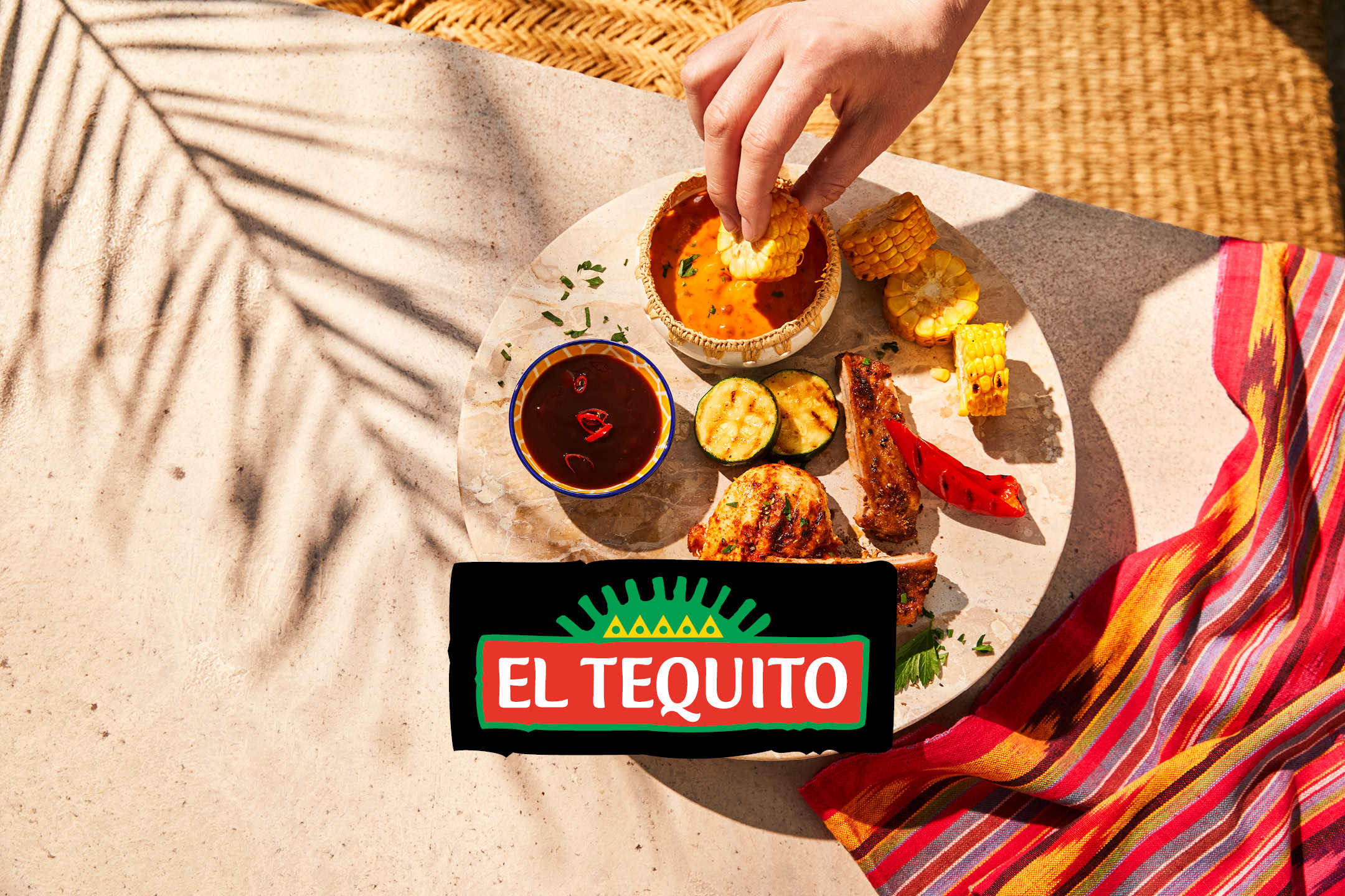 Hand dipping corn into a bowl of sauce, with grilled meat and vegetables on a platter.
