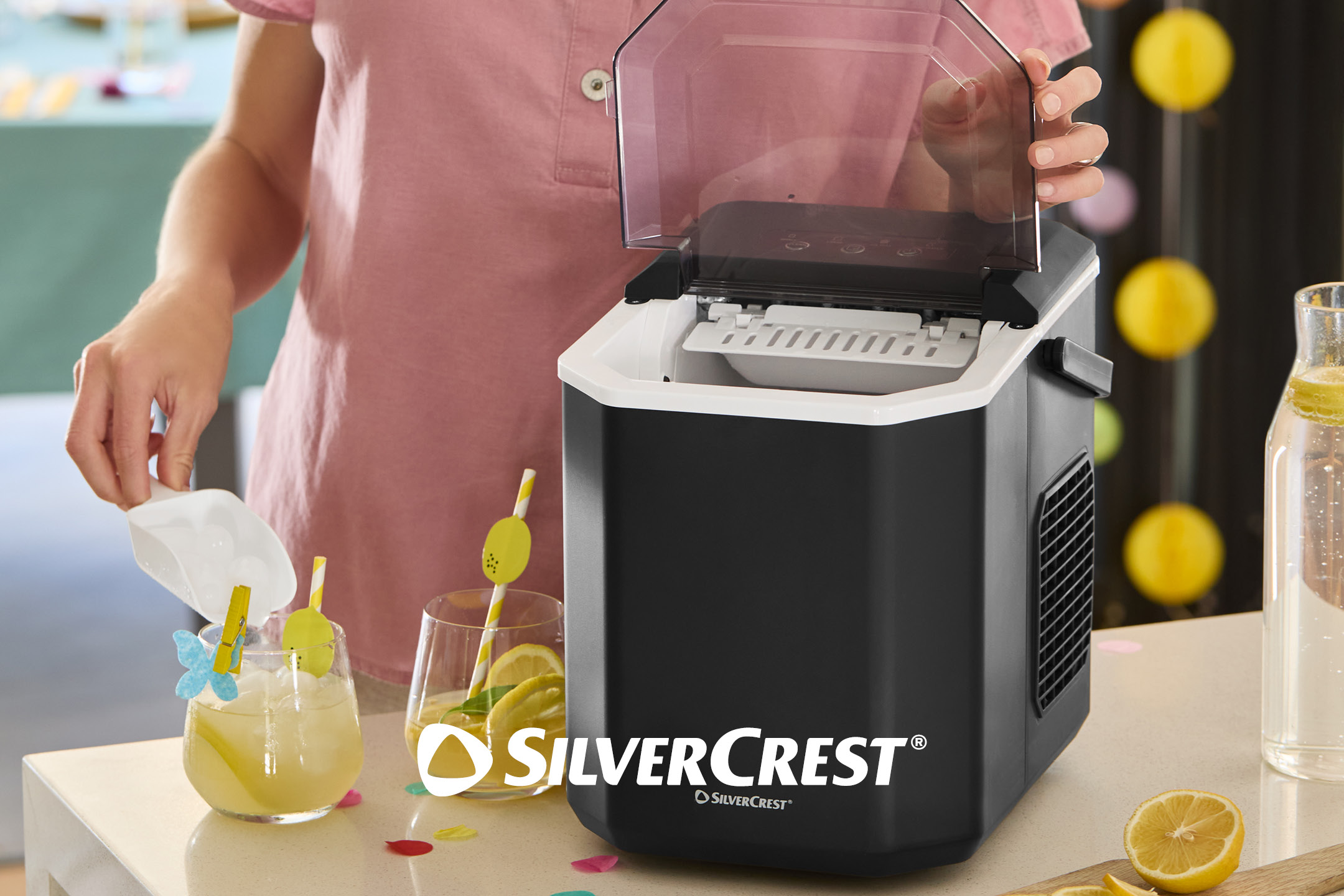 Person adding ice from an ice maker to a glass of lemonade with lemon slices.
