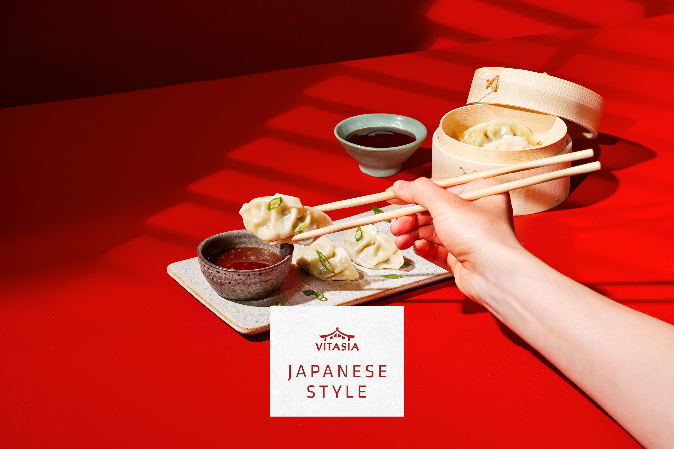 Japanese-style dumplings with dipping sauces, held by chopsticks, on a red background.