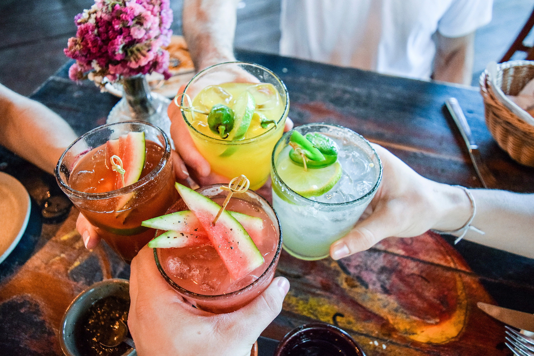 Four hands holding colorful cocktails with fruit garnishes, clinking glasses over a wooden table.