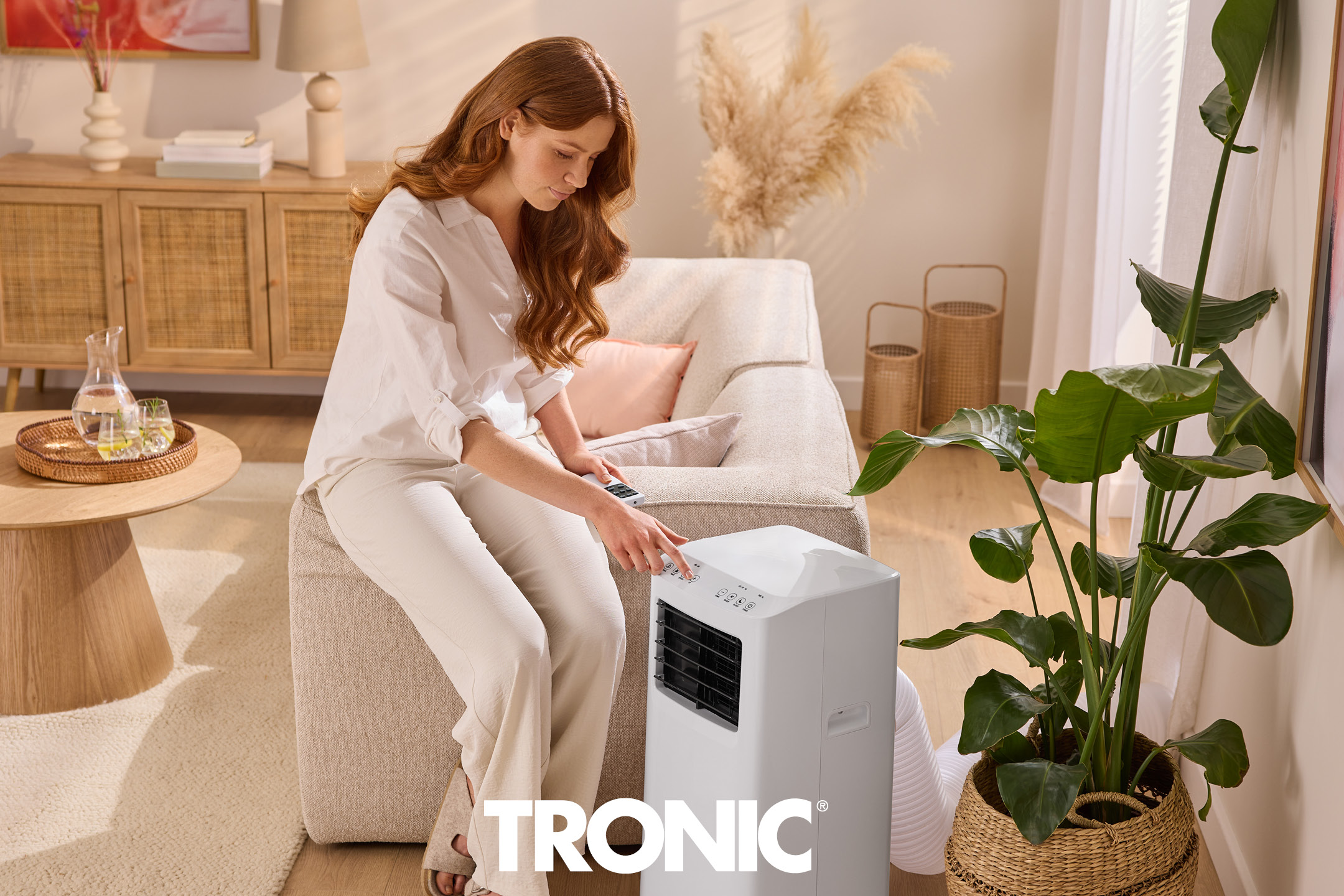 Woman on a sofa operating a portable air conditioner in a bright, modern living room.