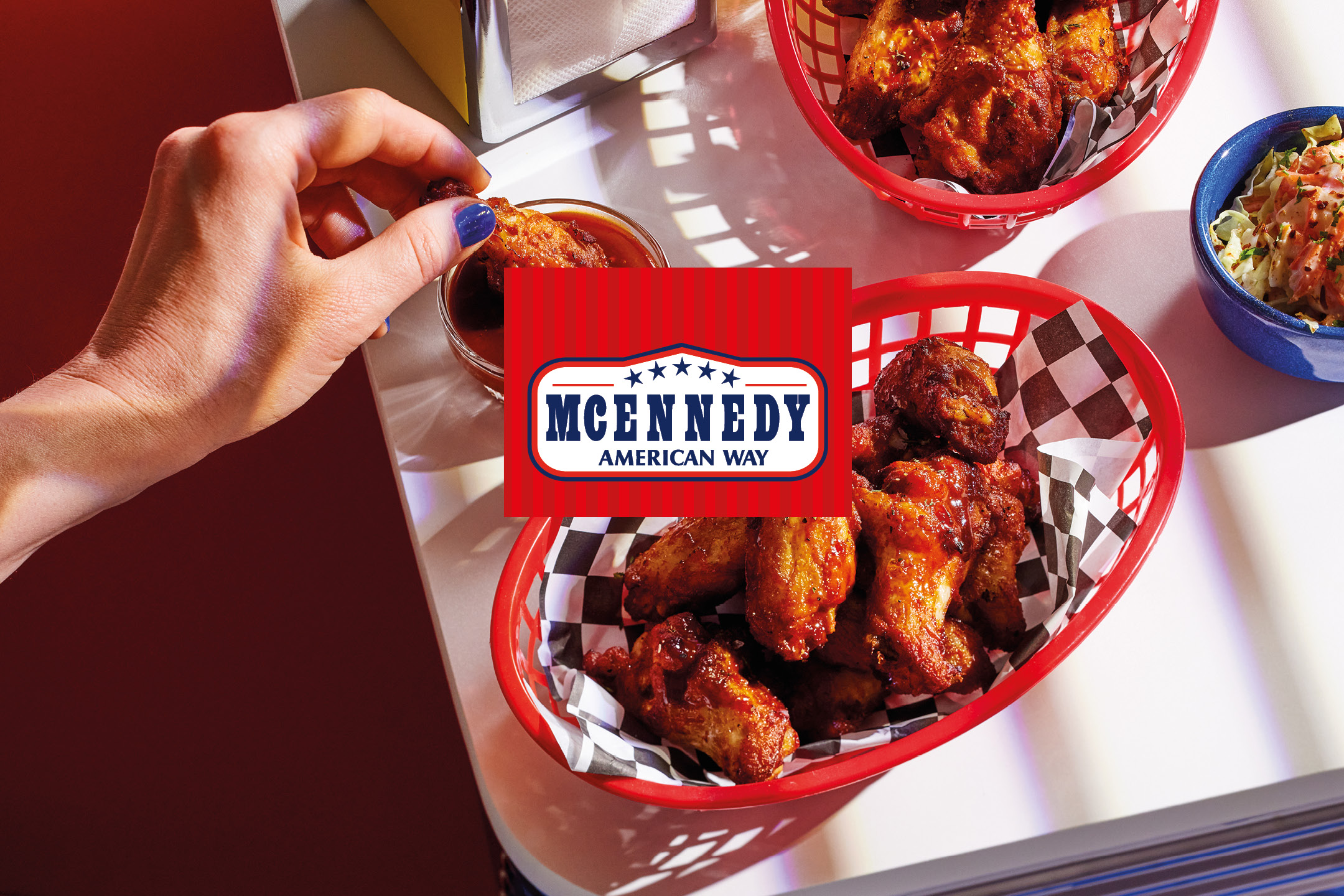 Hand dipping a chicken wing into sauce, with baskets of wings and a side dish on a table.