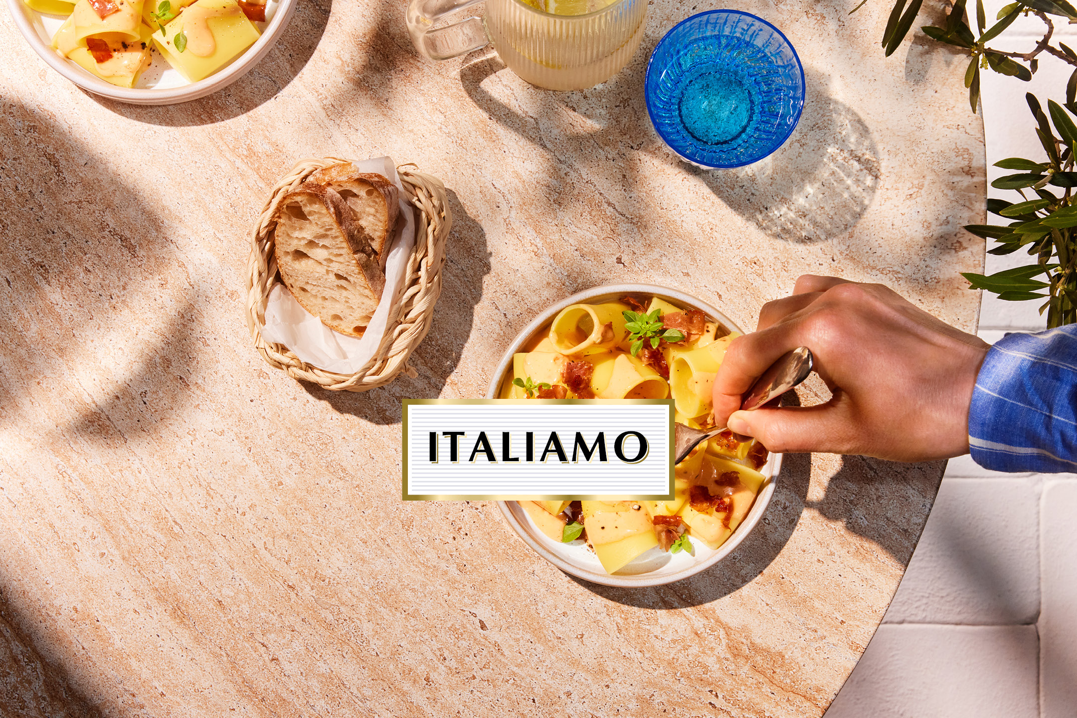 Overhead shot of pasta dishes, bread, and drinks on a table with text 'ITALIAMO'.