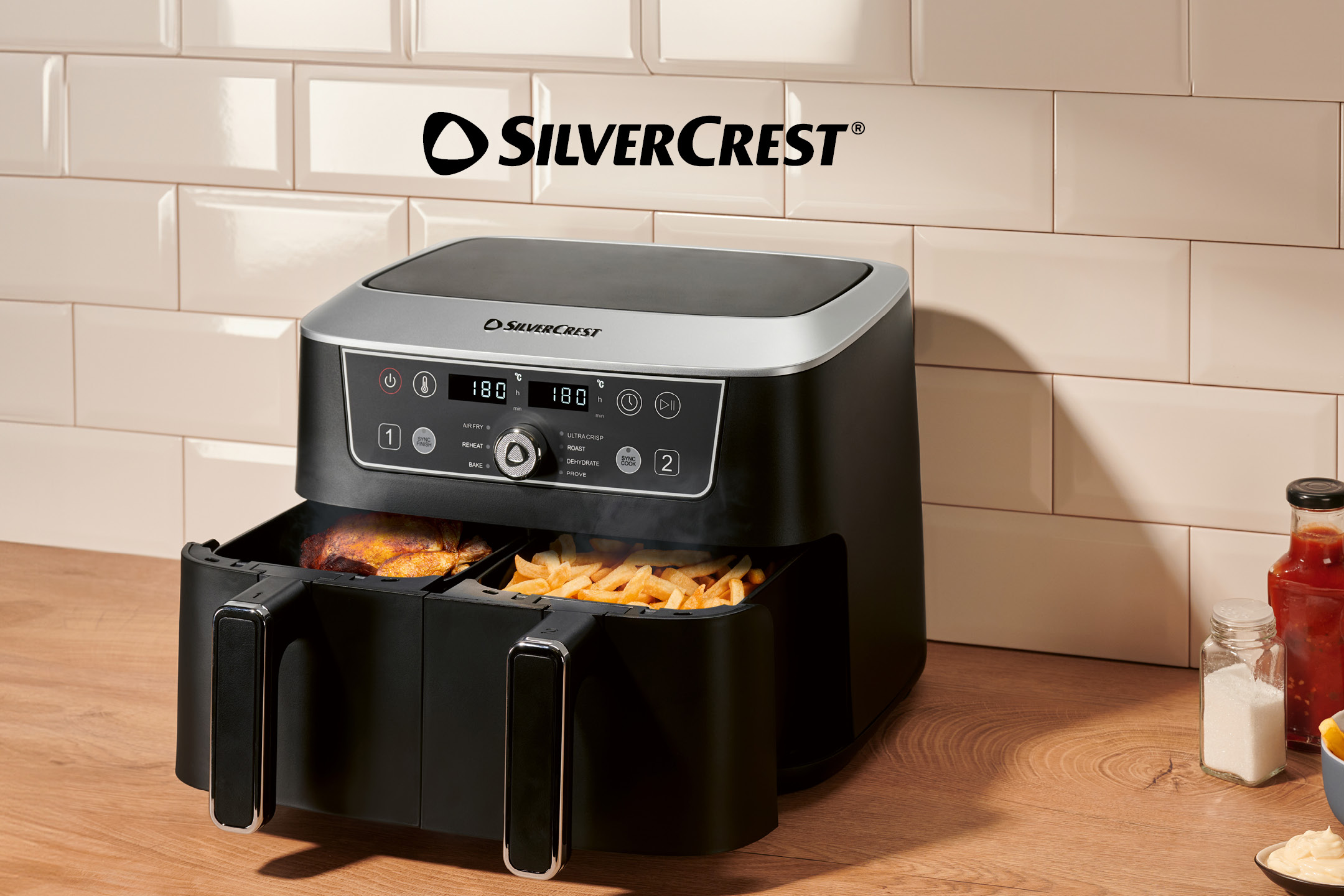 Black air fryer with two baskets, one containing chicken and the other fries, on a wooden counter.