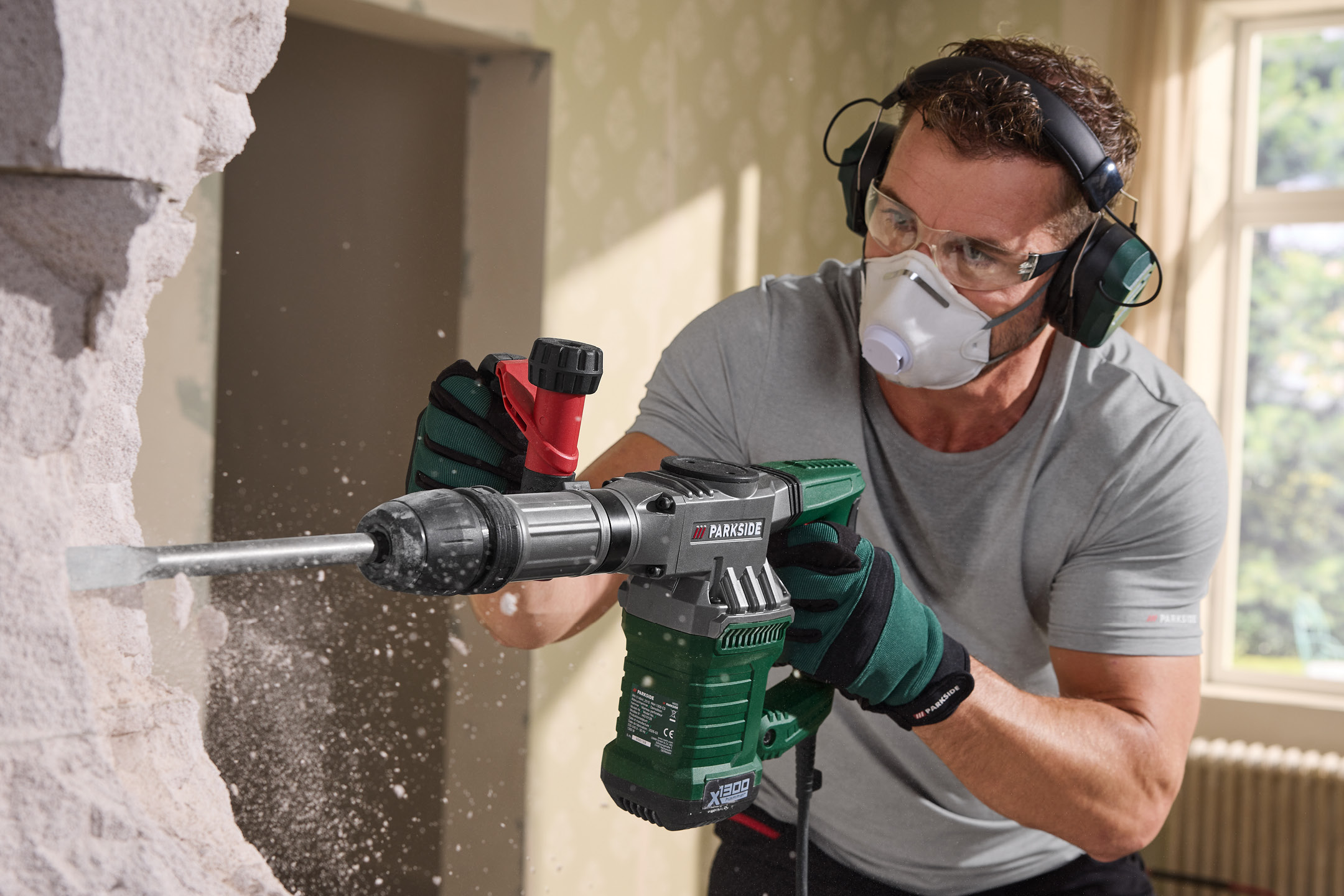 Man using a demolition hammer to break a wall, wearing safety glasses, a respirator, and ear protection.