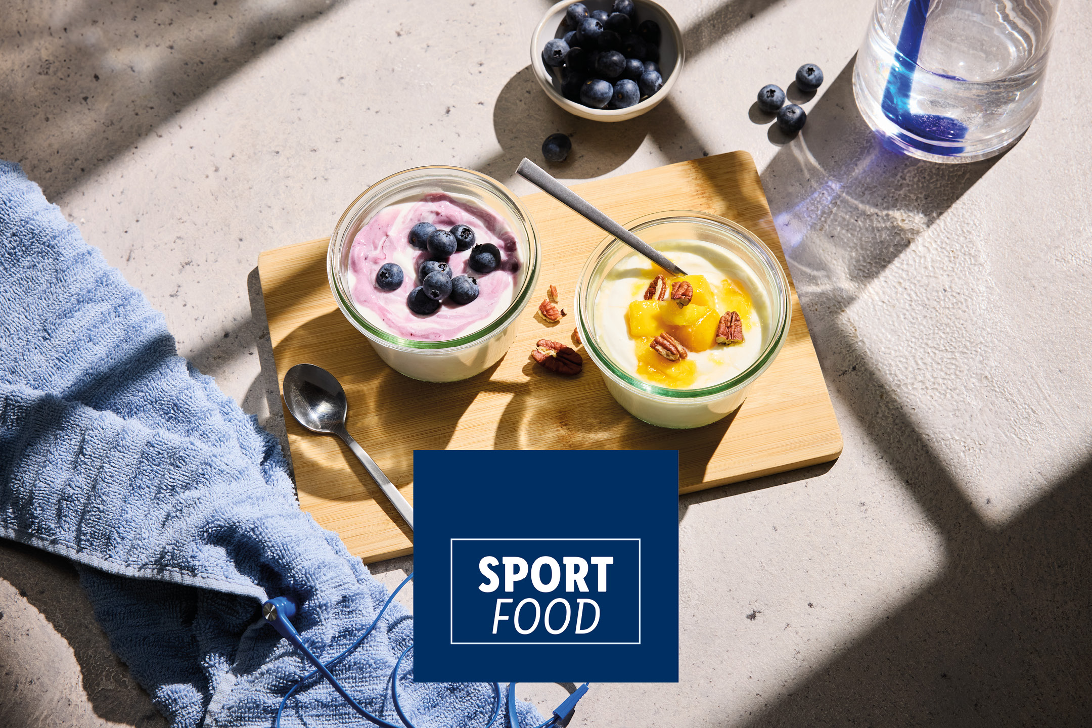 Two jars of yogurt with fruit and nuts, a bowl of blueberries, and a water bottle on a table with text 'SPORT FOOD'.