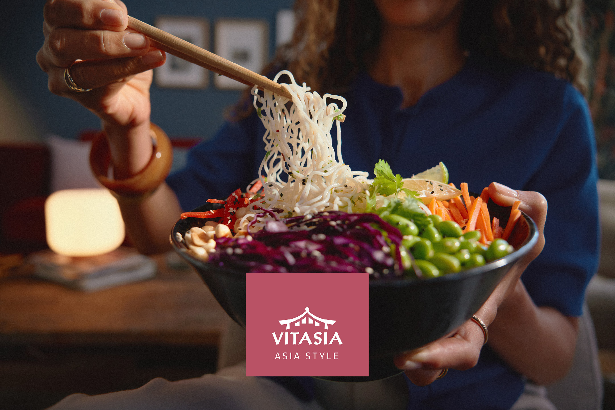 A person holding a bowl of Asian-style noodles with vegetables, lifting some noodles with chopsticks.