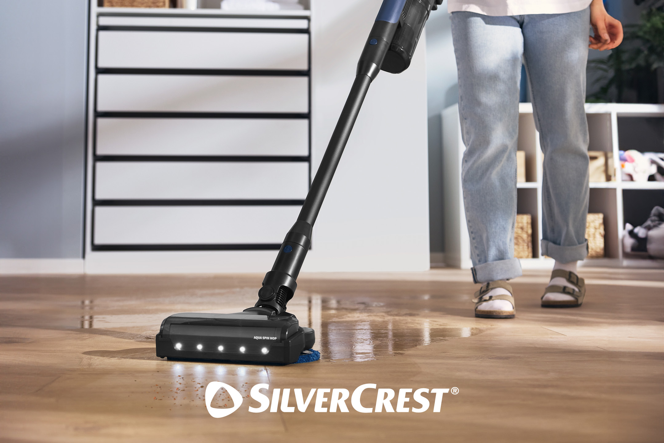 A floor cleaner with LED lights cleaning a wet wooden floor, with a person in sandals nearby.