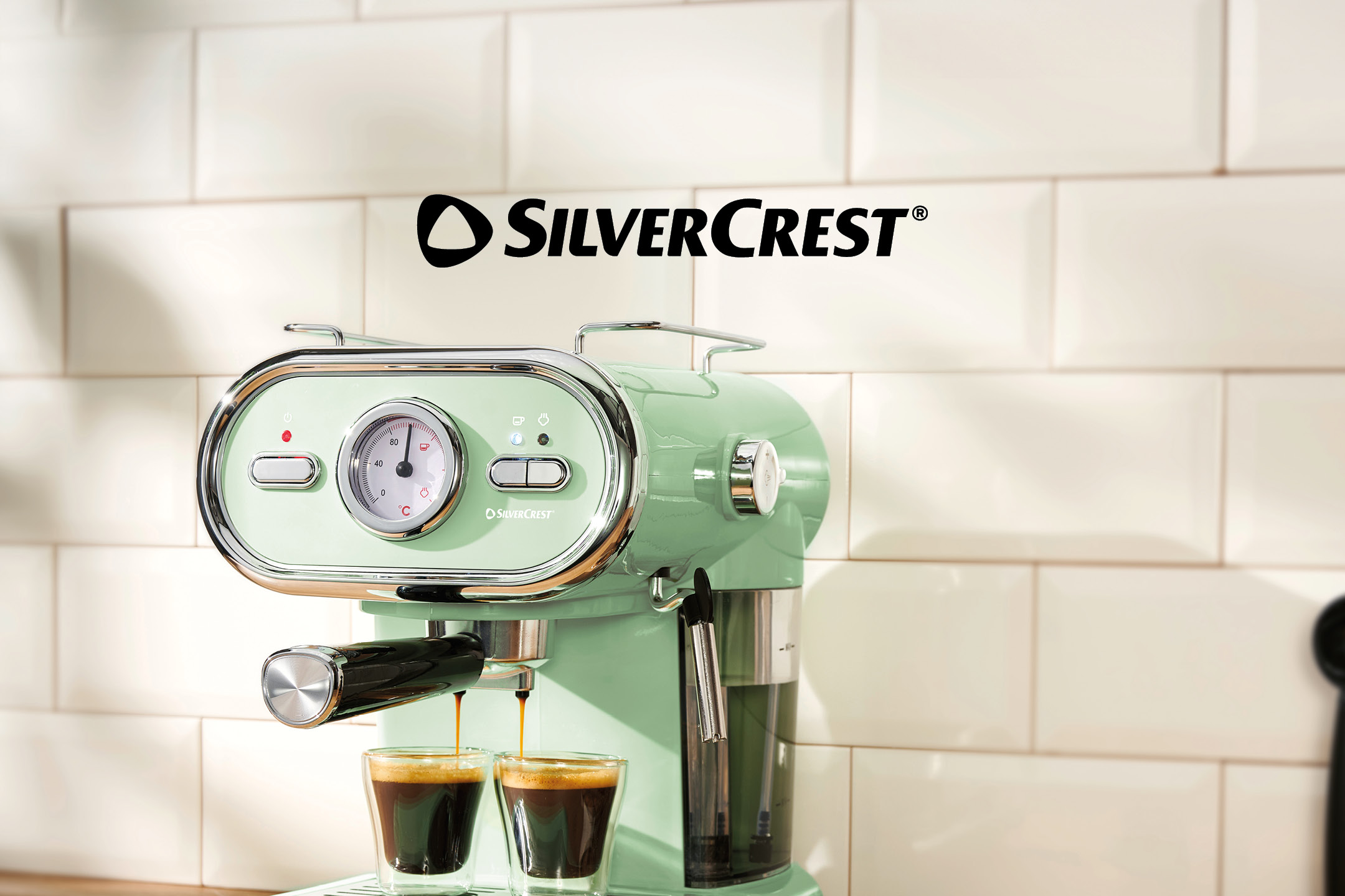 Mint green espresso machine brewing coffee into two small glasses on a tiled kitchen wall.