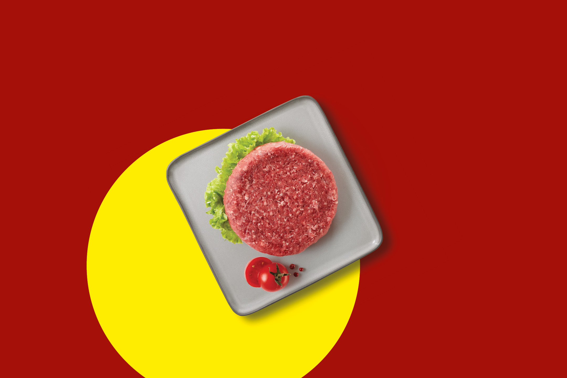 Raw beef burger patty with lettuce and tomatoes on a grey plate, on a red and yellow background.