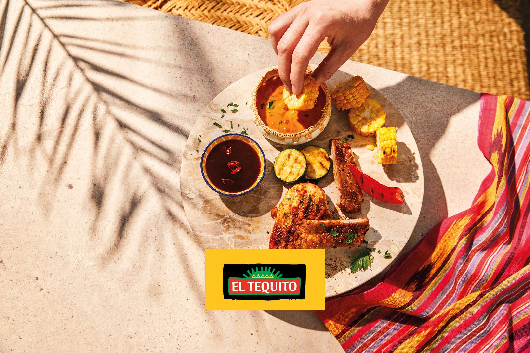 Grilled ribs, chicken, corn, zucchini, and sauces on a stone board with a colorful textile.