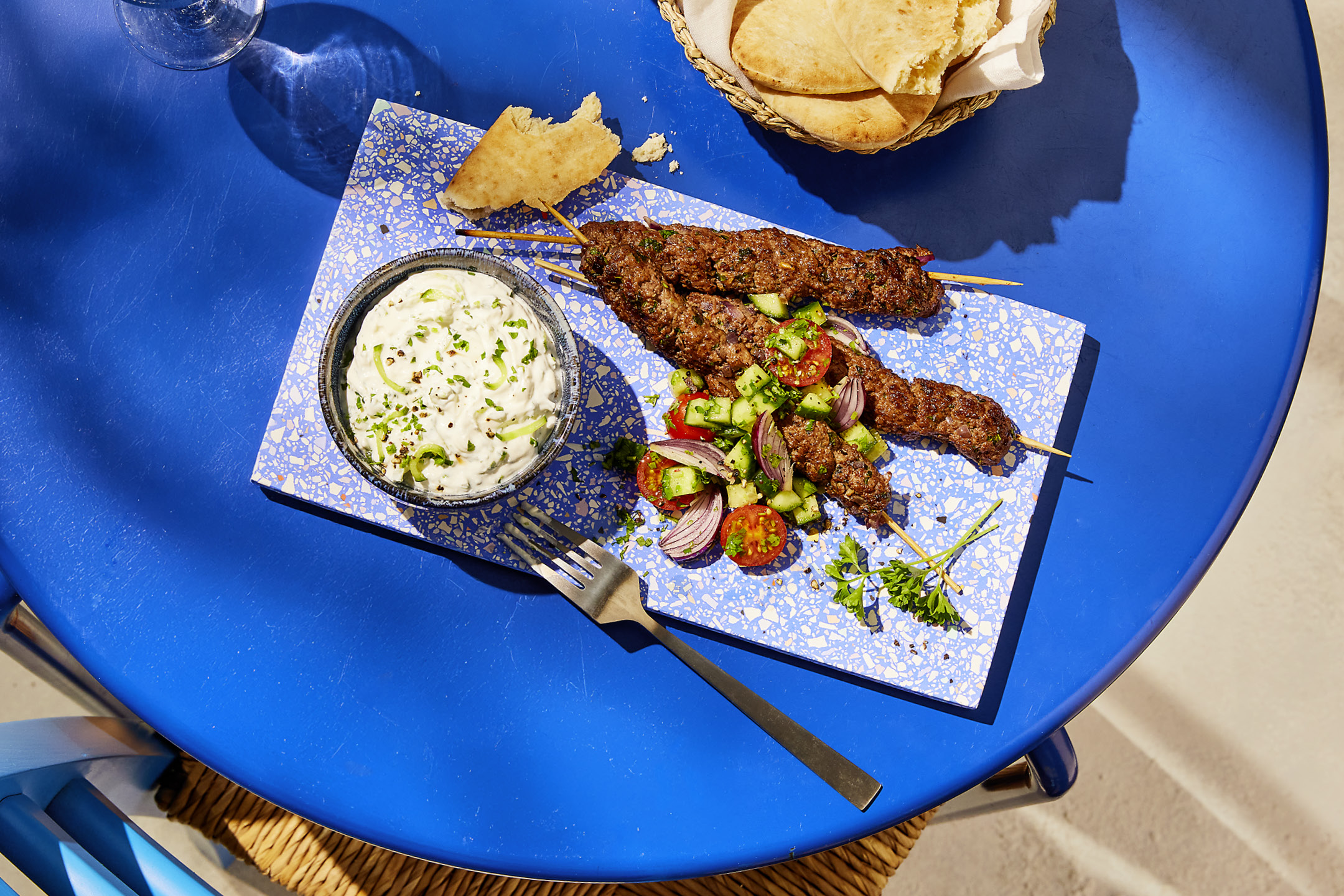Kebab skewers with a side of tzatziki and fresh salad on a blue terrazzo tray.