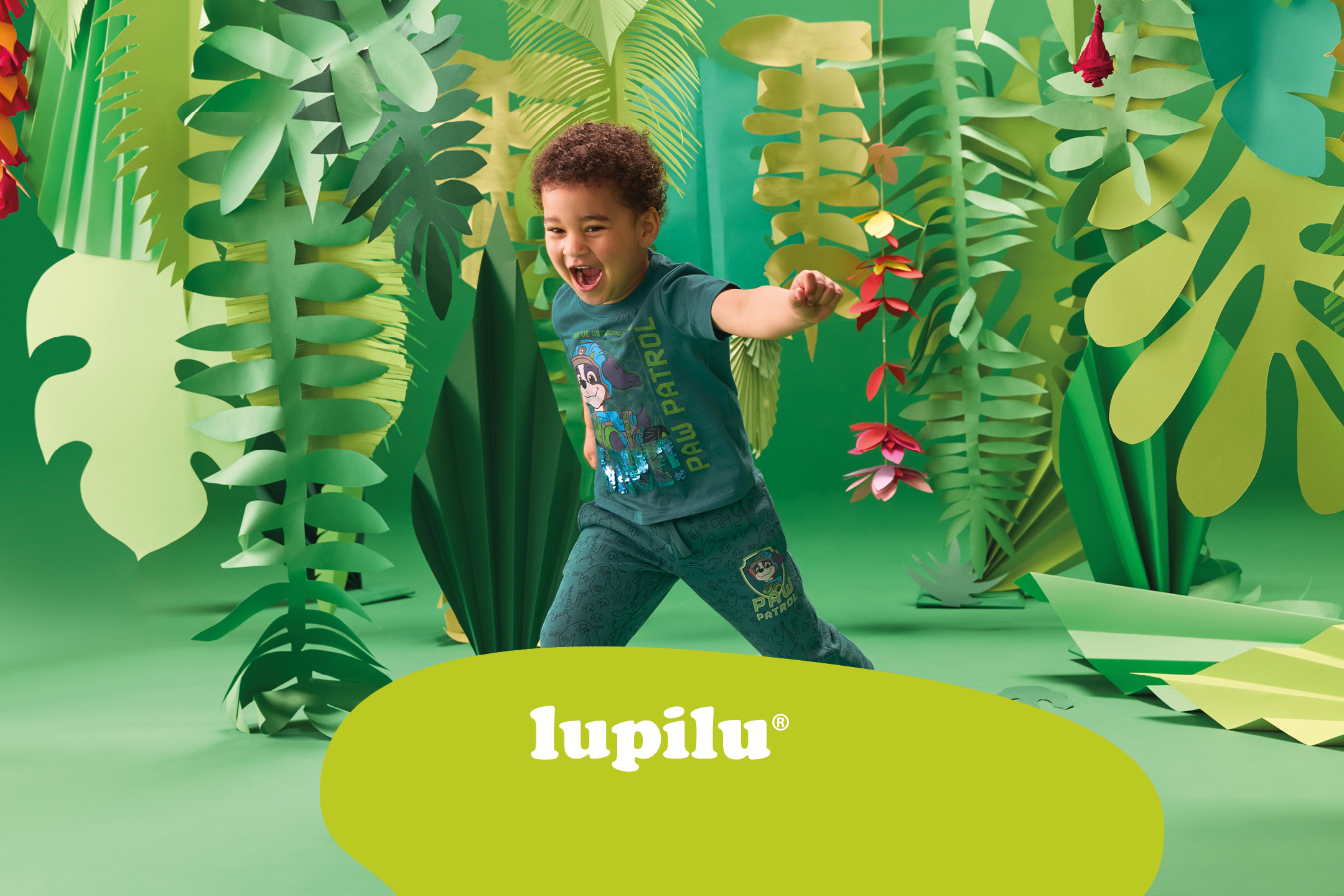 A boy in a green t-shirt with a Paw Patrol print, surrounded by paper jungle leaves.